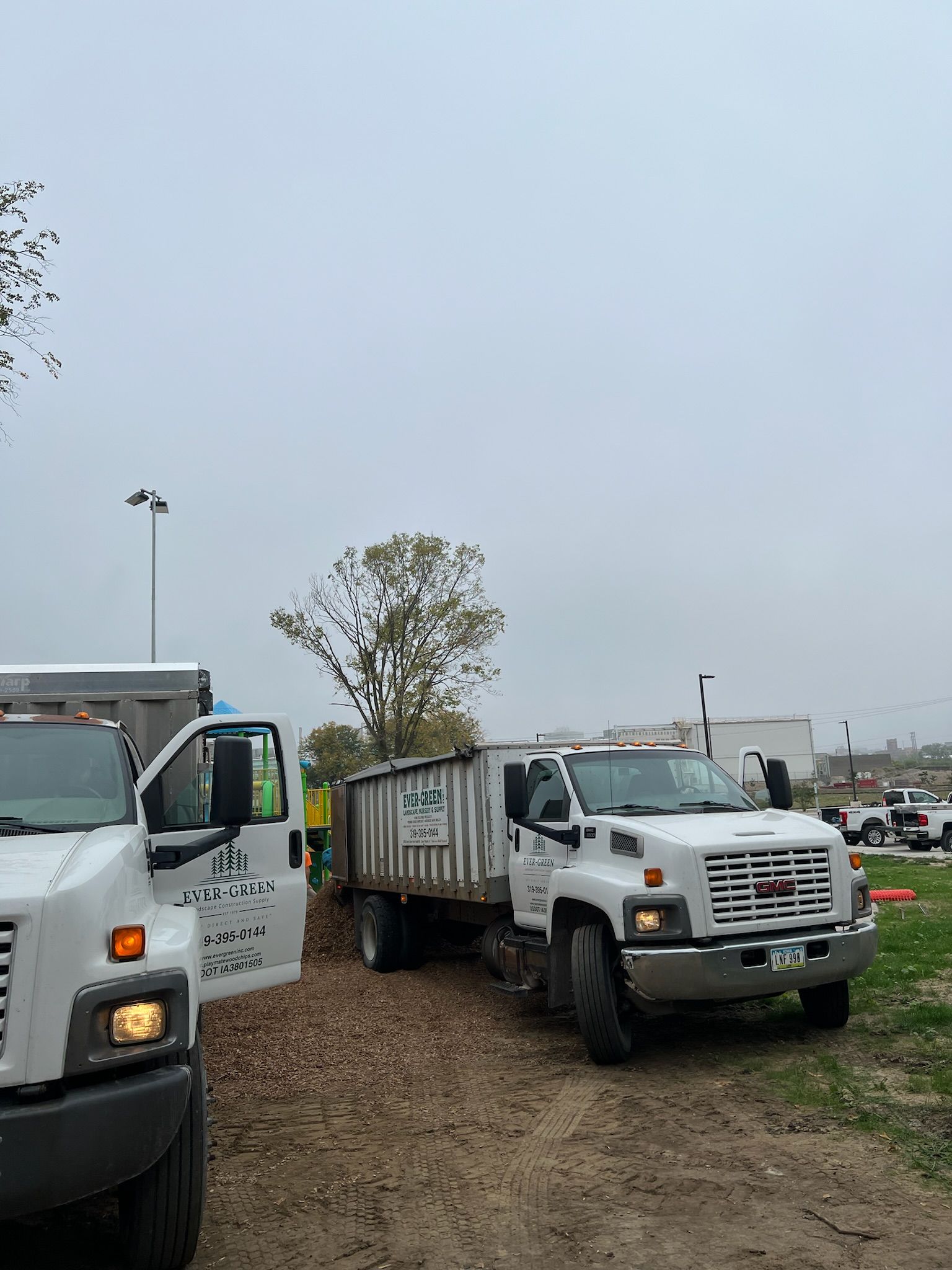 Two white dump trucks parked on dirt under an overcast sky.