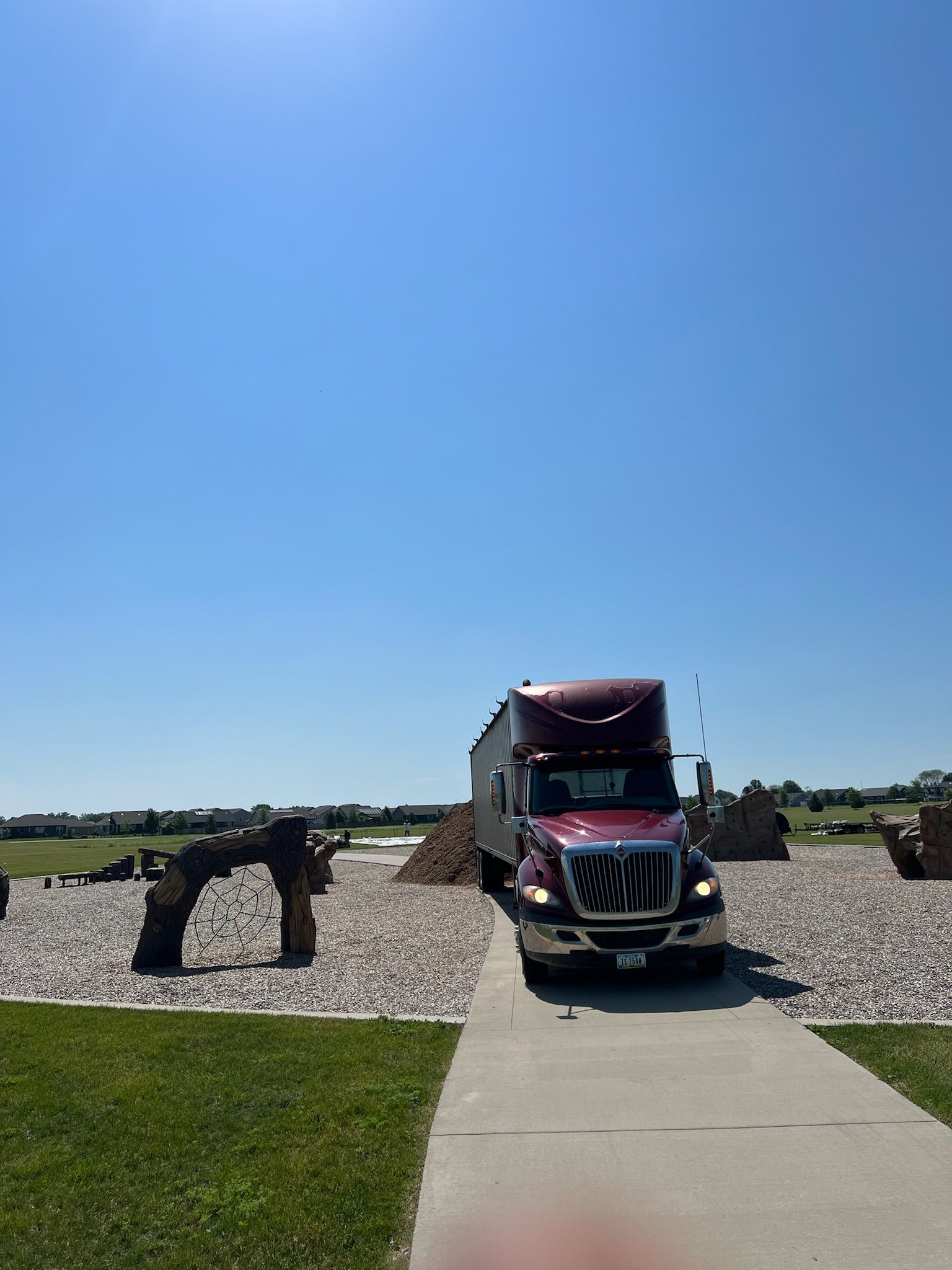 Red semi-truck parked on a concrete path; clear blue sky, grassy area, and rock feature in background.