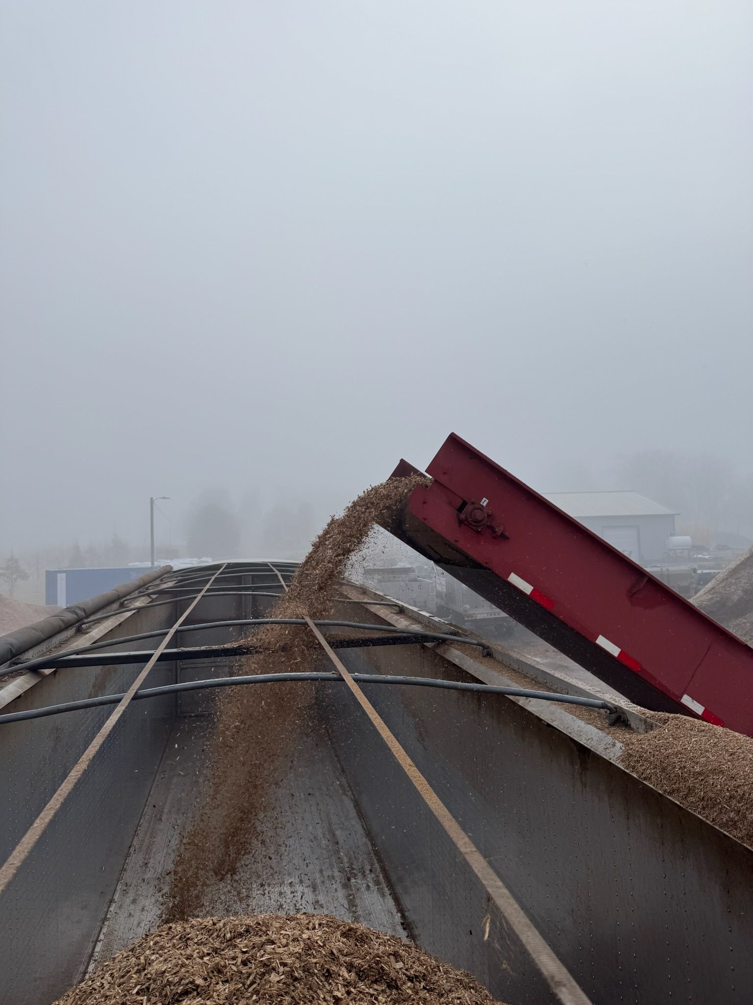Brown gravel pouring from a red conveyor belt onto a flatbed truck in a foggy outdoor setting.