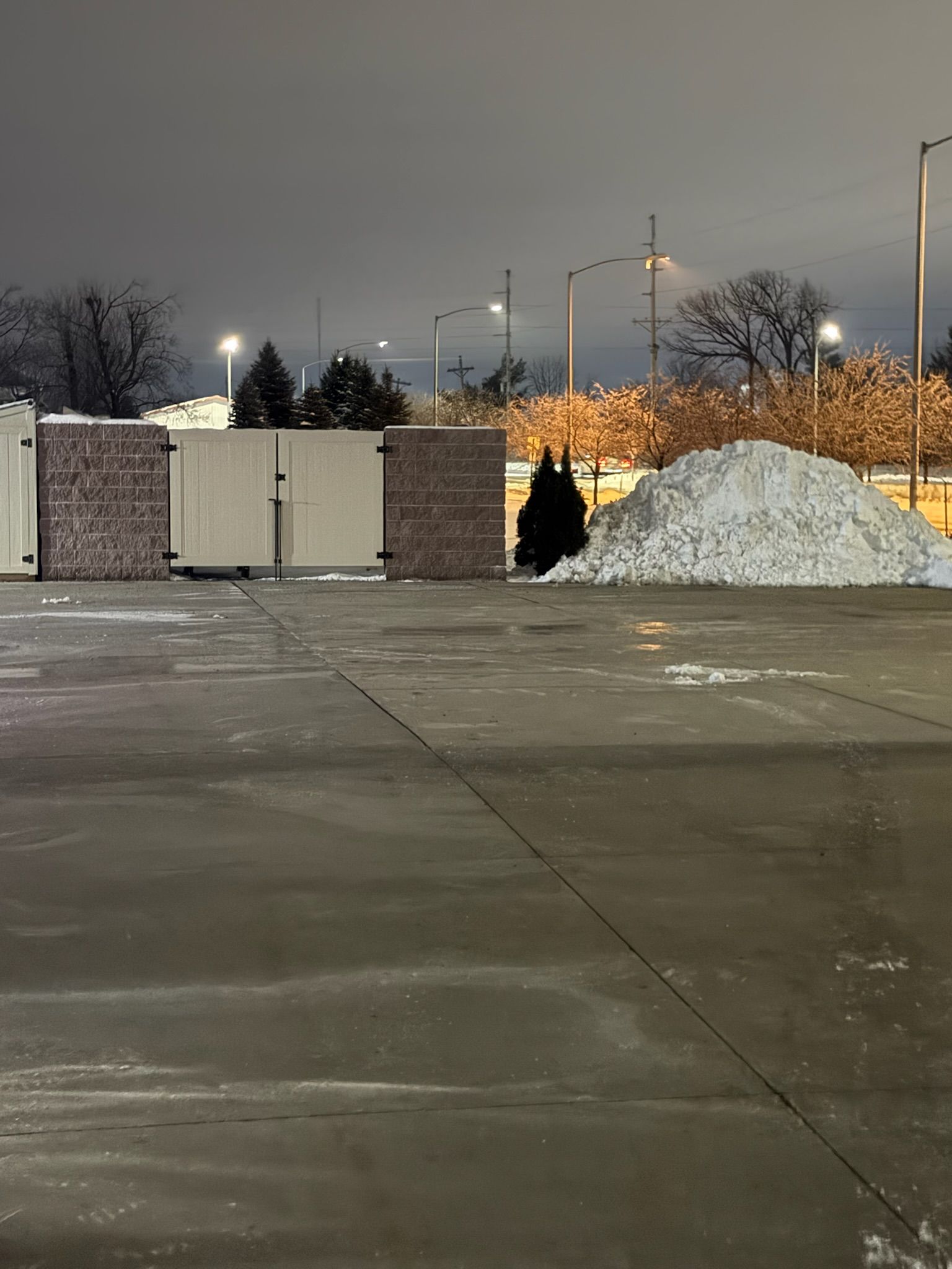 Nighttime scene: snowy pavement in front of beige structures and a pile of snow. Streetlights glow.
