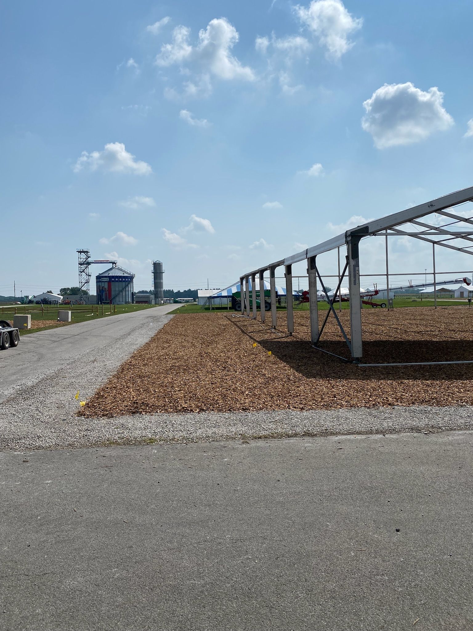 Gravel road leads past a structure's frame and a gravel bed toward farm buildings under a blue sky.