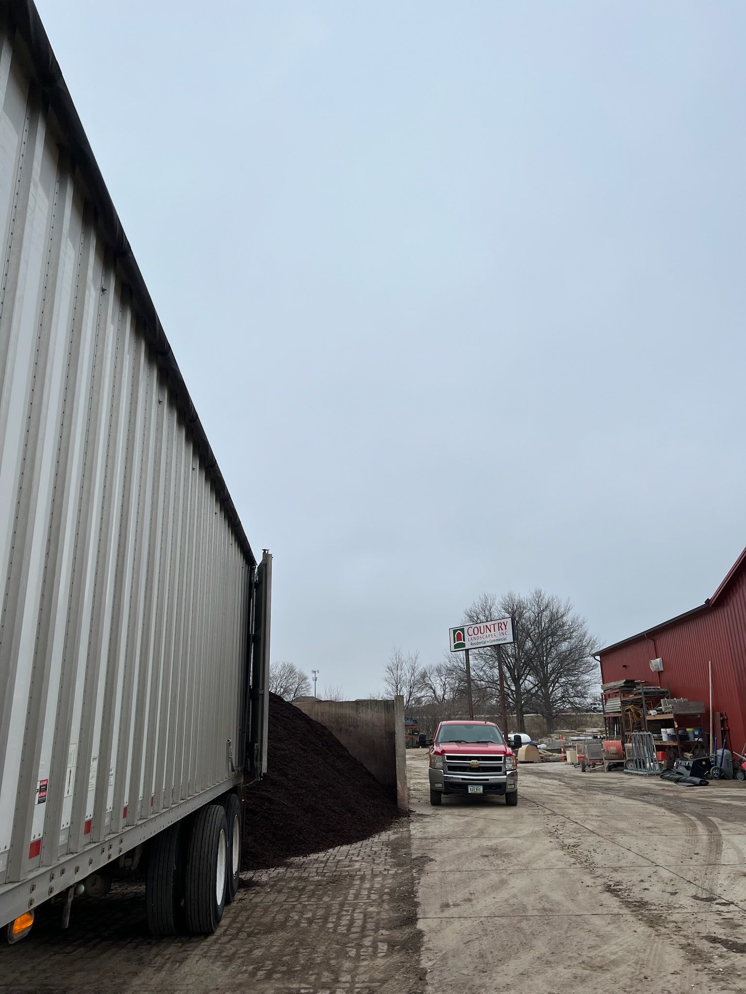 Truck parked near a large pile of dark material, with a red truck and building visible under a cloudy sky.