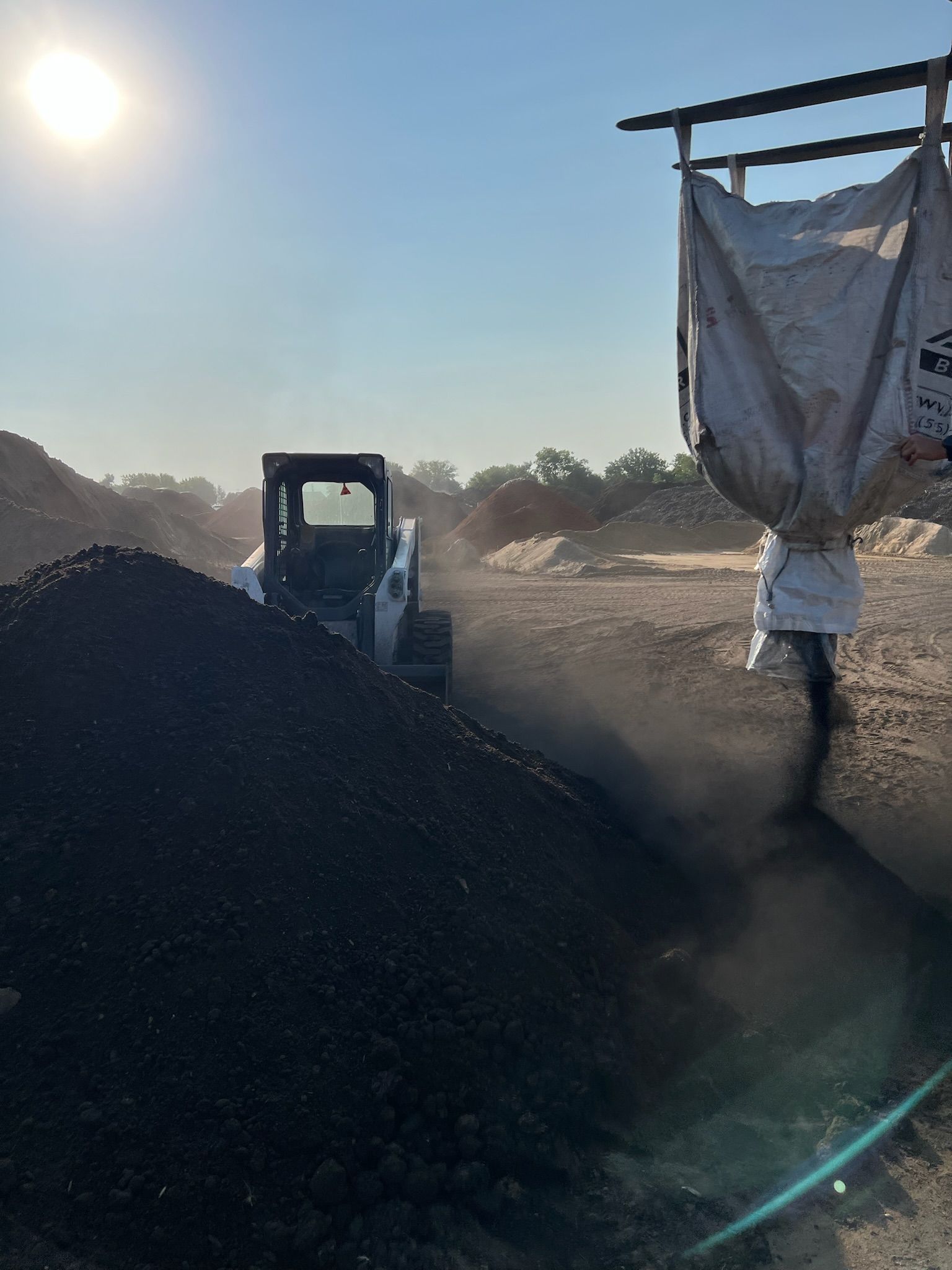 A skid steer loader moving dark material under a large suspended bag, bright sunlight overhead.