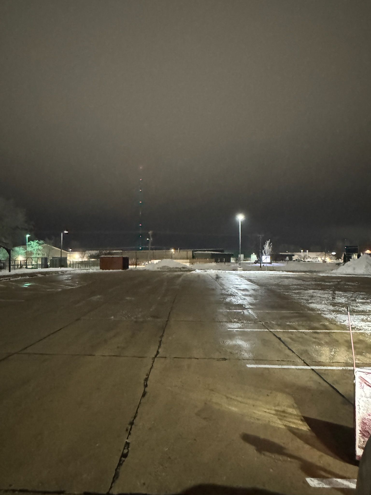 Nighttime view of an empty parking lot, snow-covered, with distant lights and a radio tower. Overcast sky.