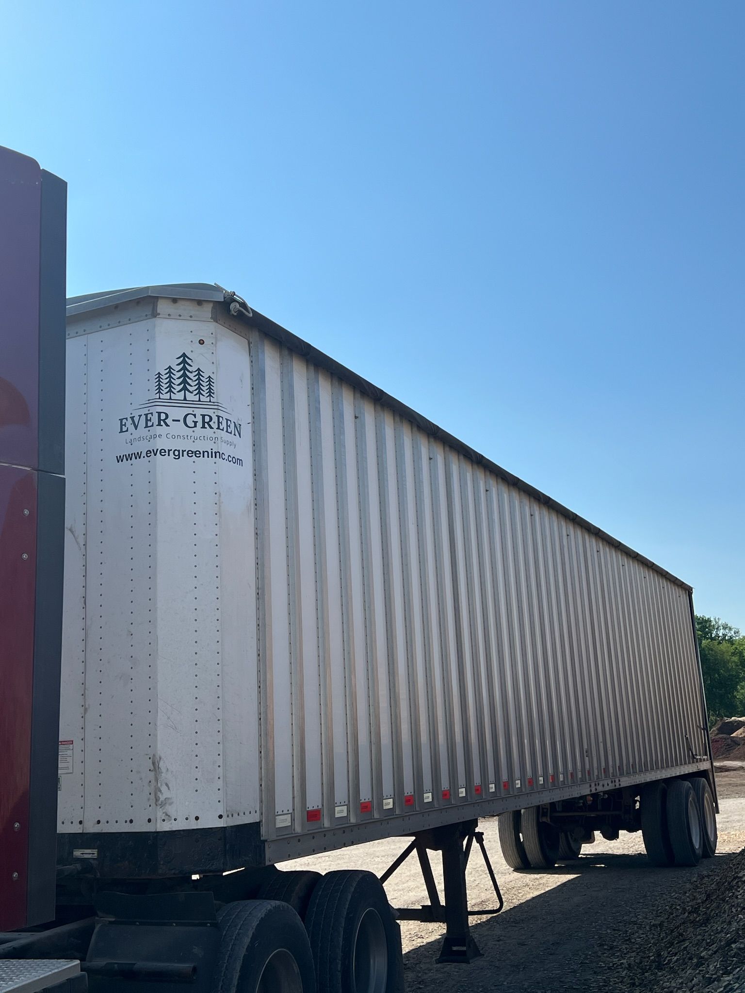 Semi-truck trailer with white corrugated sides, parked on gravel against a clear blue sky.