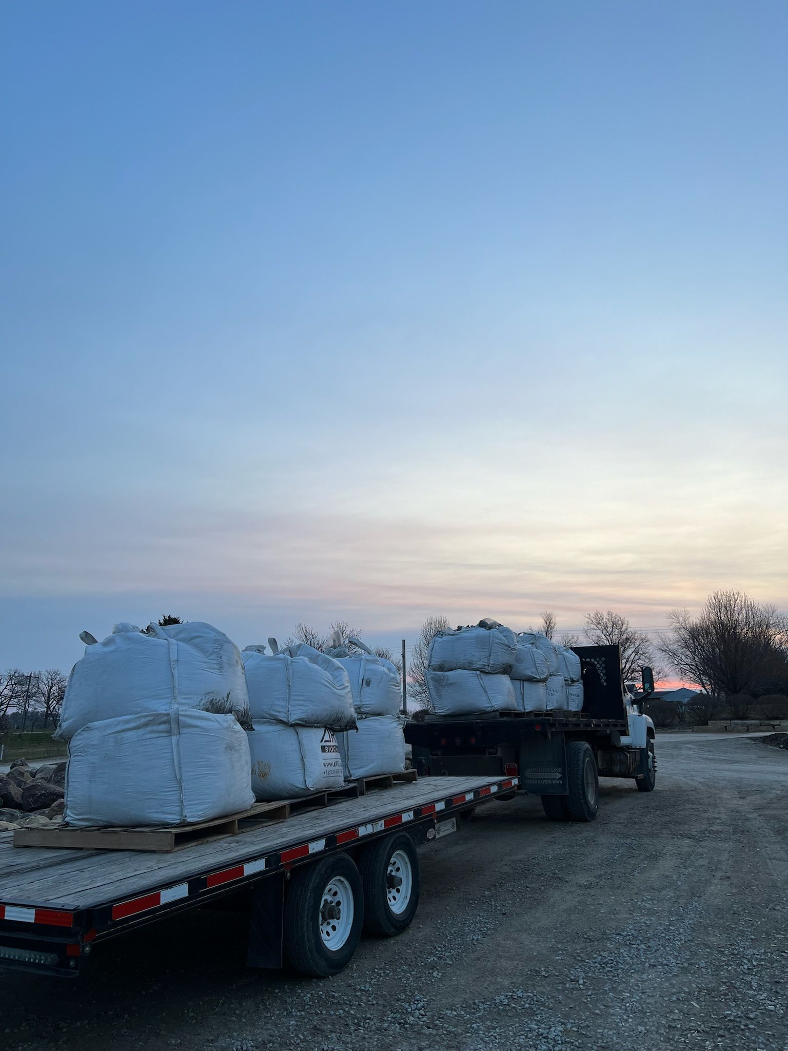Truck hauling a flatbed trailer loaded with large, white sacks against a twilight sky.