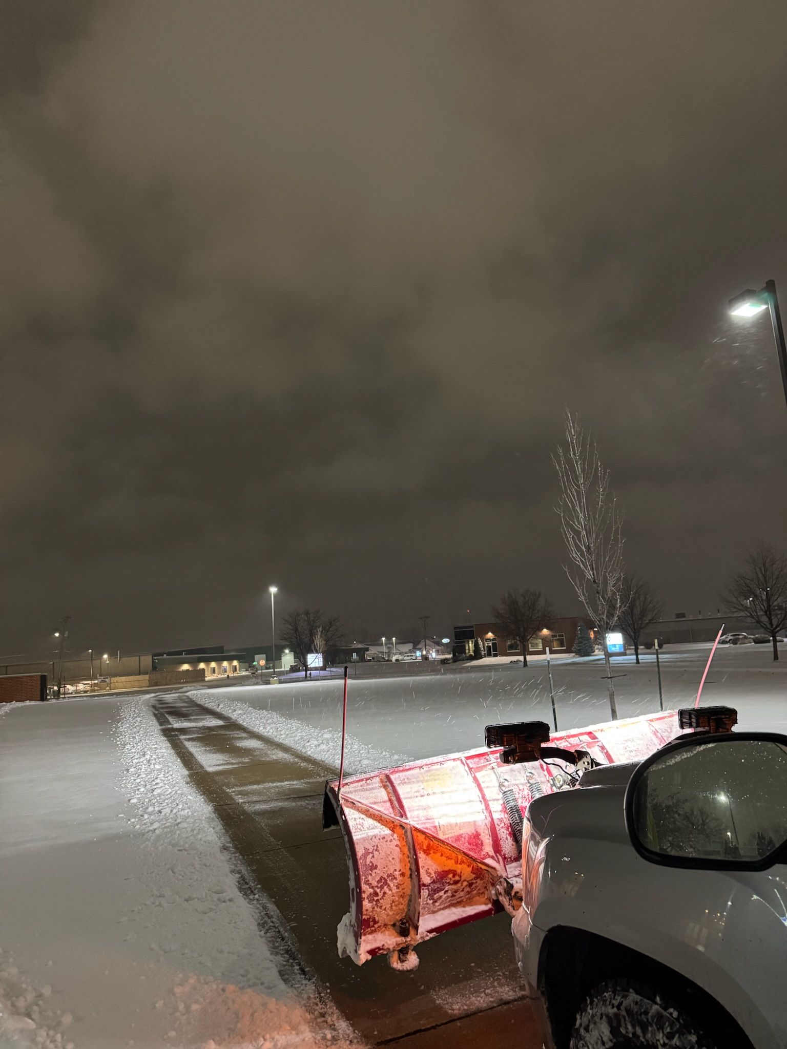 Snowplow clearing a snow-covered sidewalk at night. White snow contrasts with the dark sky.