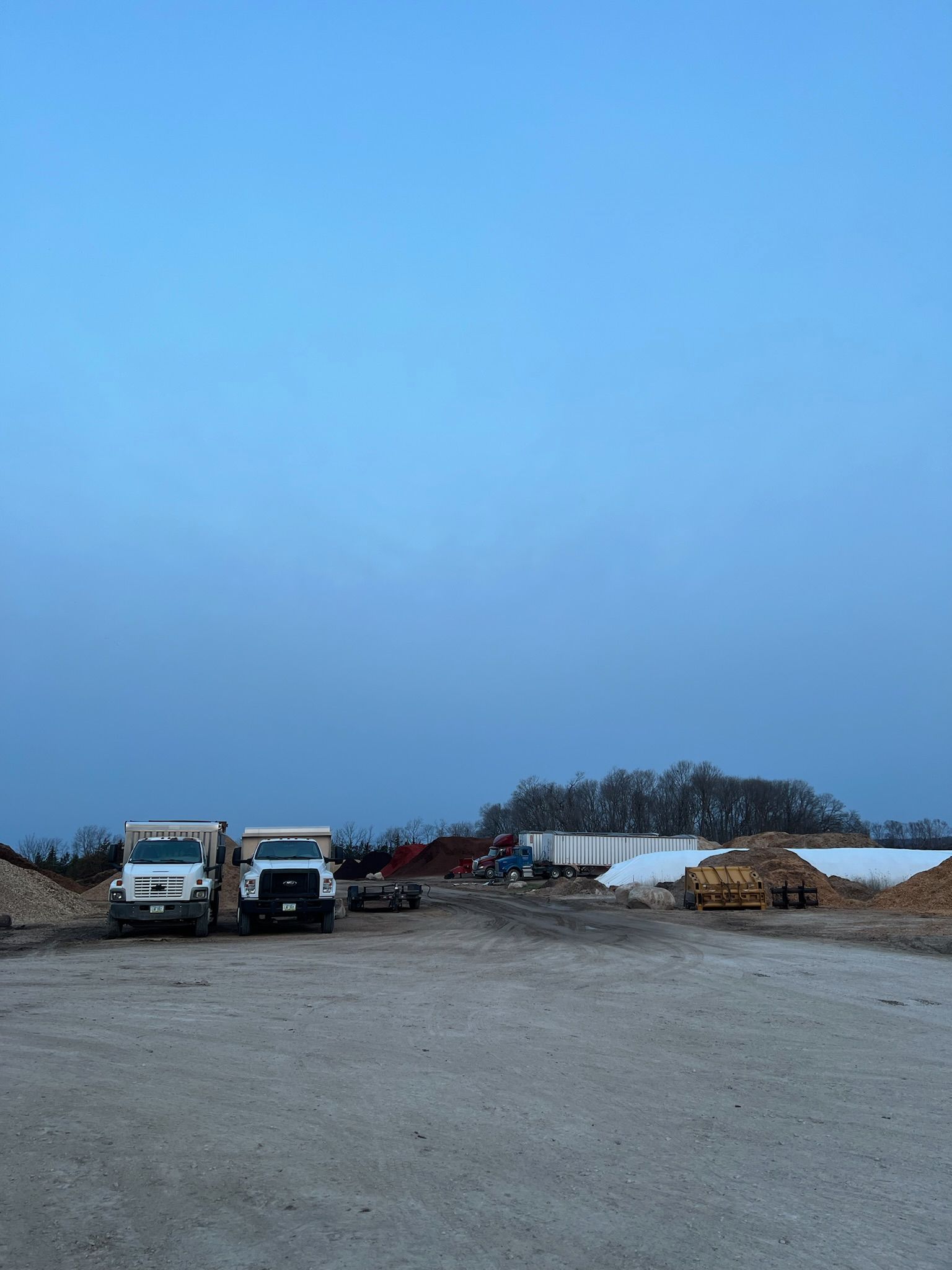 Trucks parked on a gravel lot under a pale blue sky near piles of wood and trees.