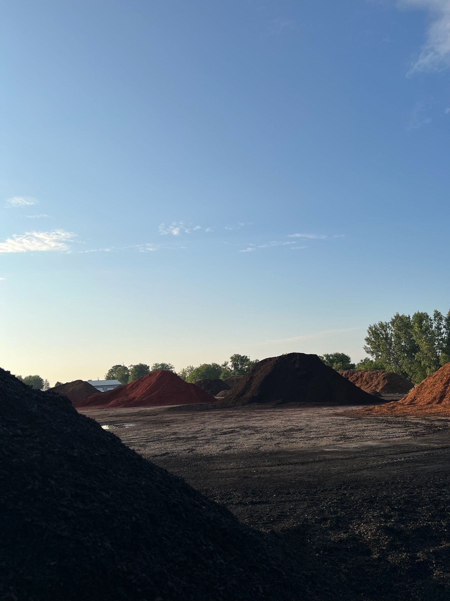 Piles of mulch in various colors under a blue sky, trees in the background.