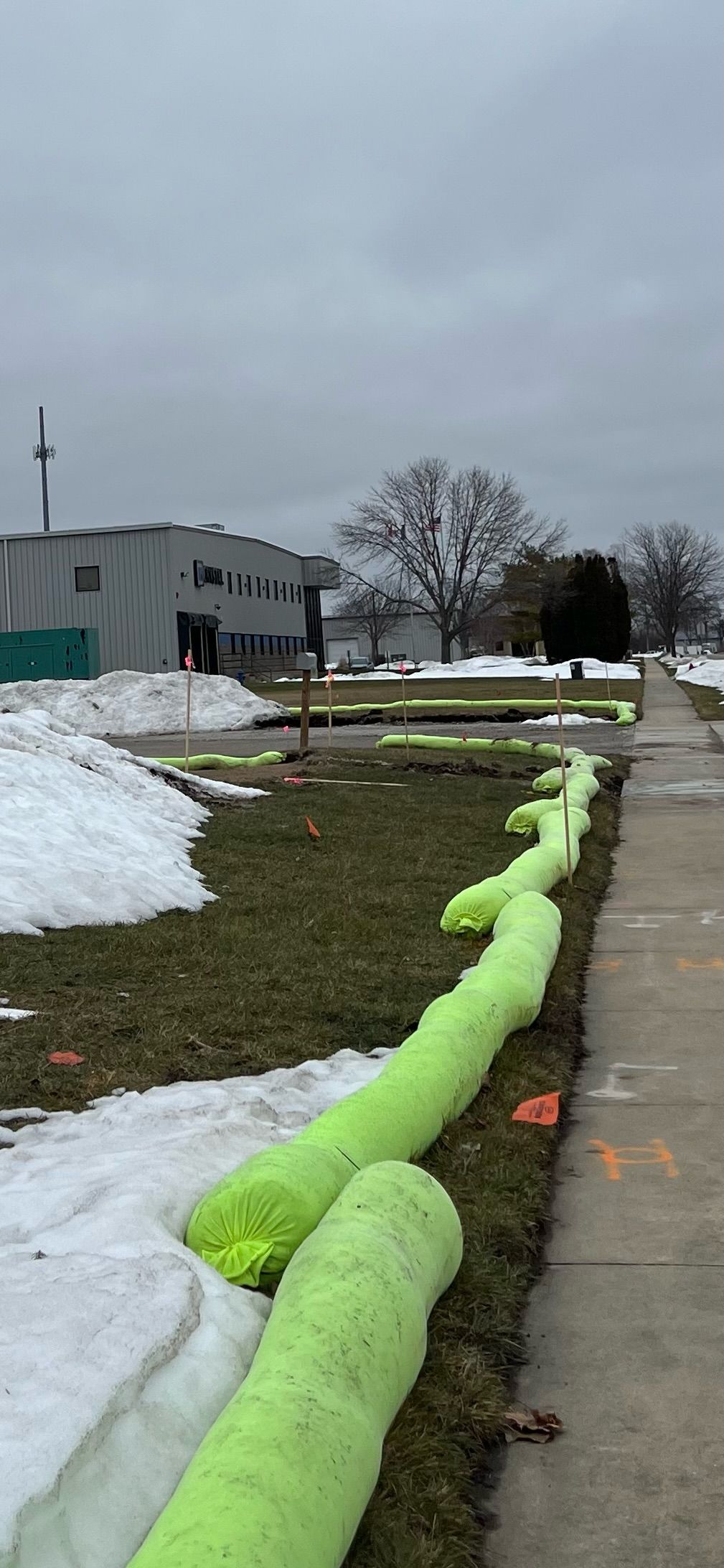 Neon-green absorbent booms line a grassy area with patches of snow next to a sidewalk and building under a gray sky.
