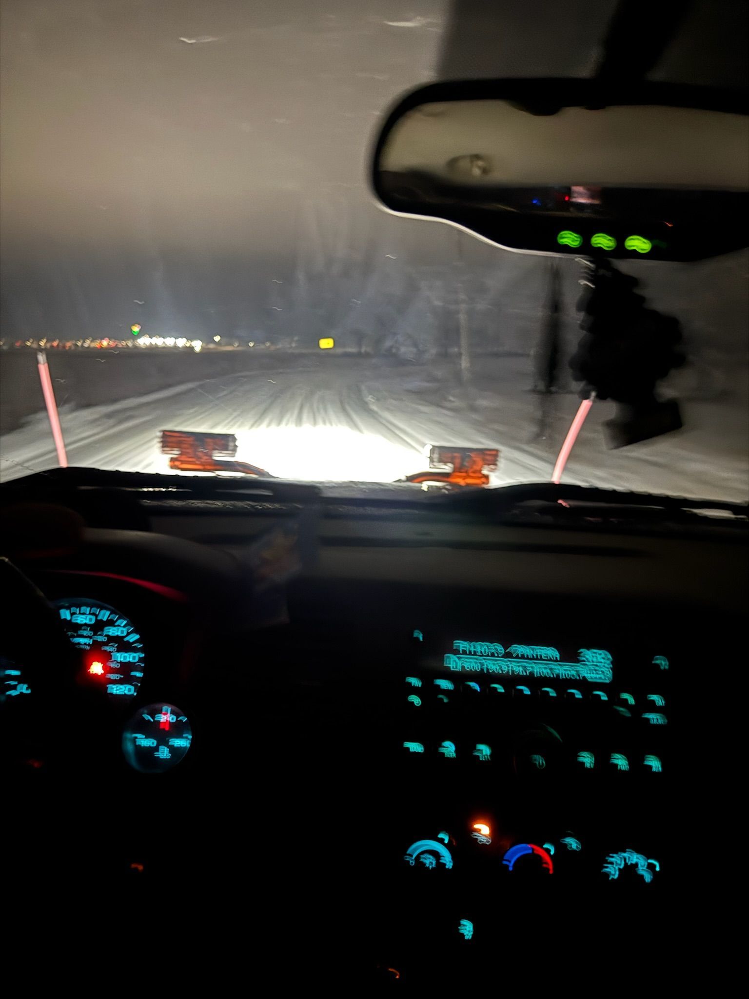 Interior view of a vehicle driving on a snowy road at night. Dashboard and road are visible with headlights on.