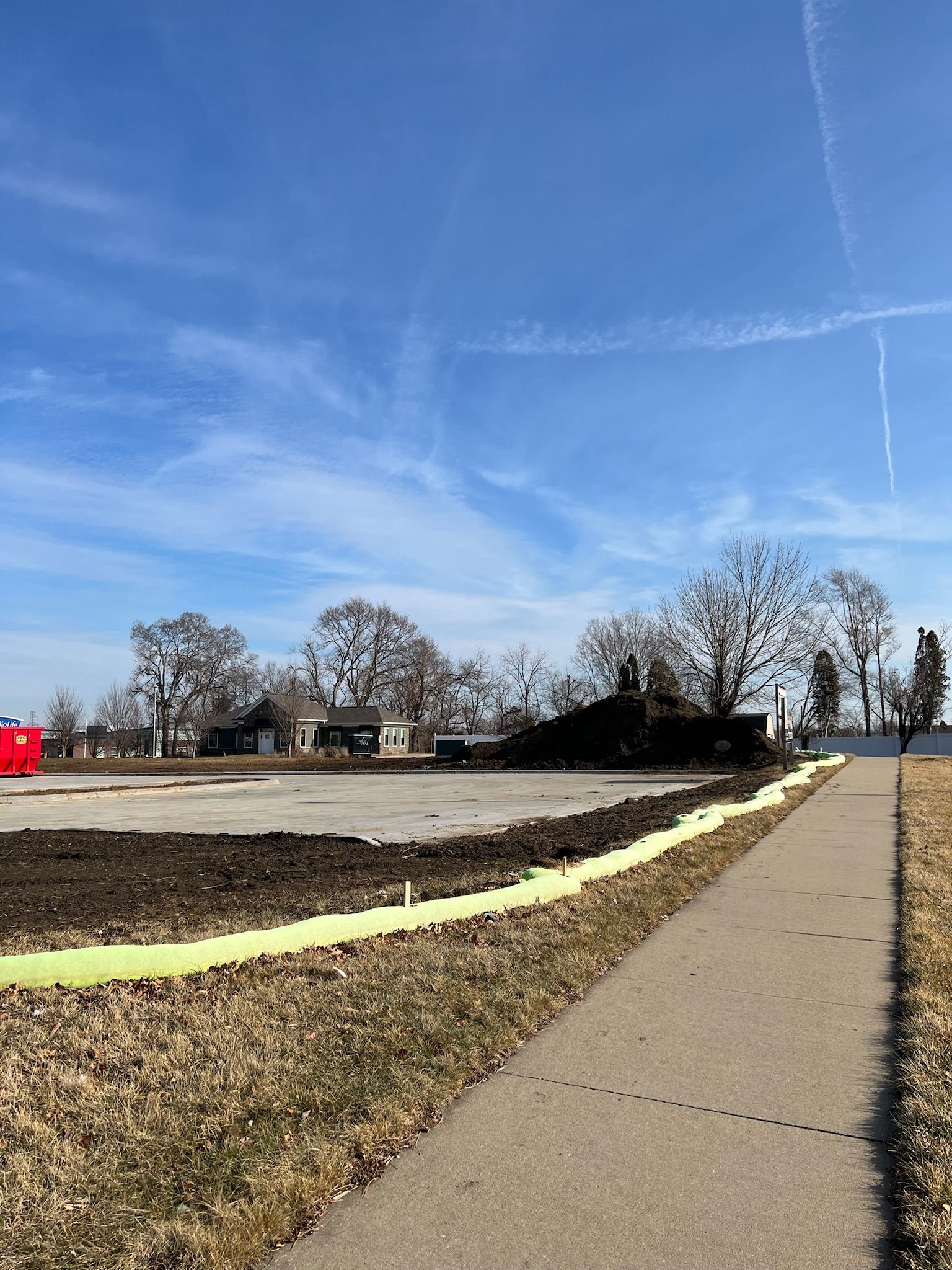 Cleared construction site next to a sidewalk, with trees and a blue sky in the background.