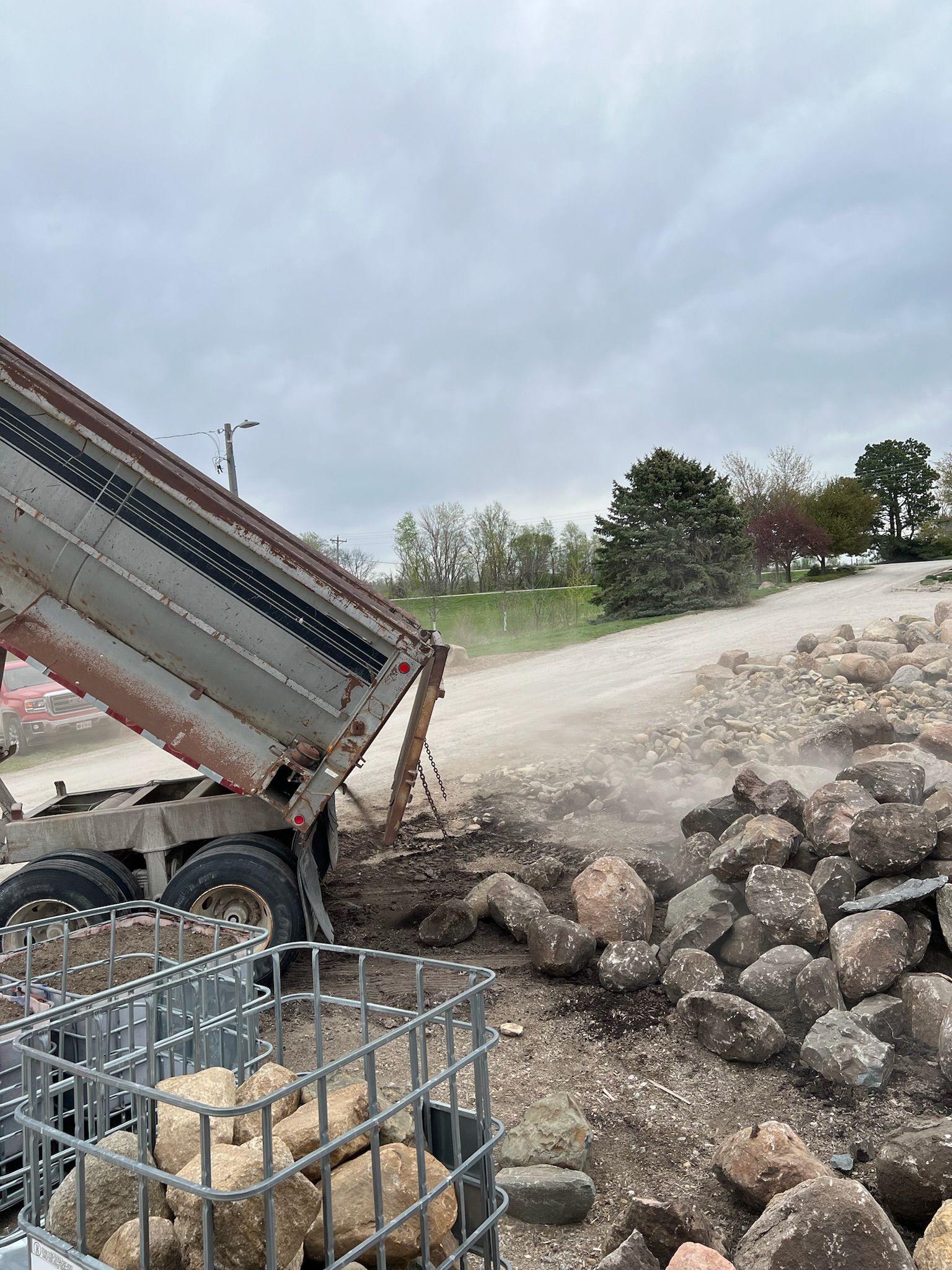 A dump truck unloads rocks onto a pile. Steel cage holds rocks in the foreground, cloudy sky overhead.