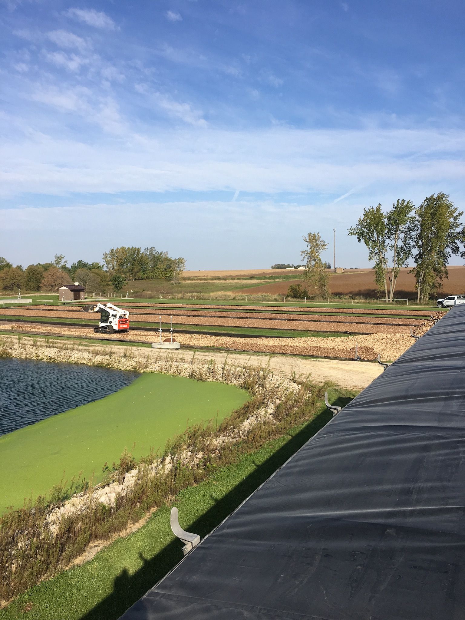 Overhead view of a field with a pond. Construction equipment and plots with various color soils are visible. Blue sky.