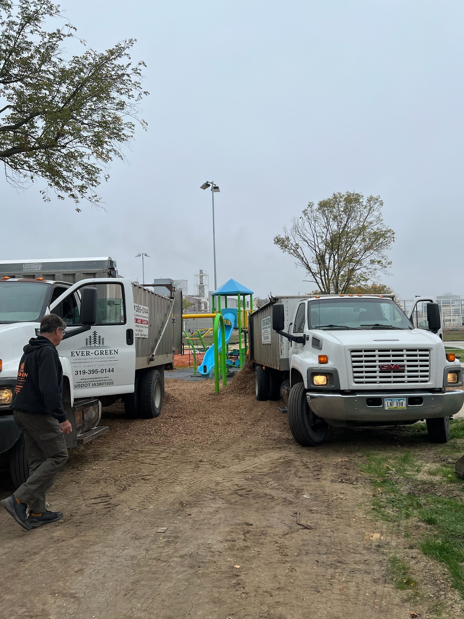 Two dump trucks removing leaves from a park path near a playground on an overcast day.
