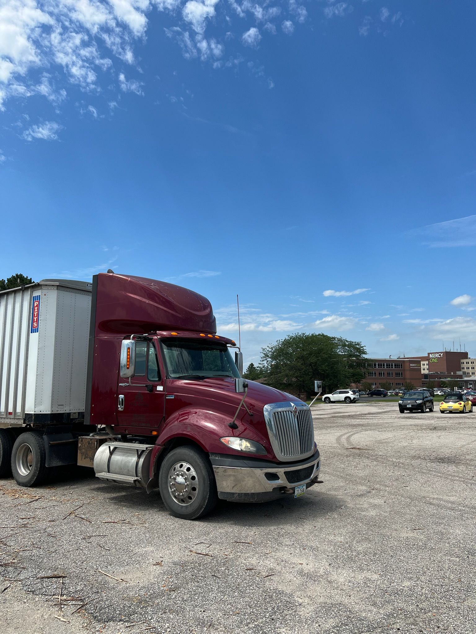 Maroon semi-truck with white trailer parked on a gravel lot under a blue sky.