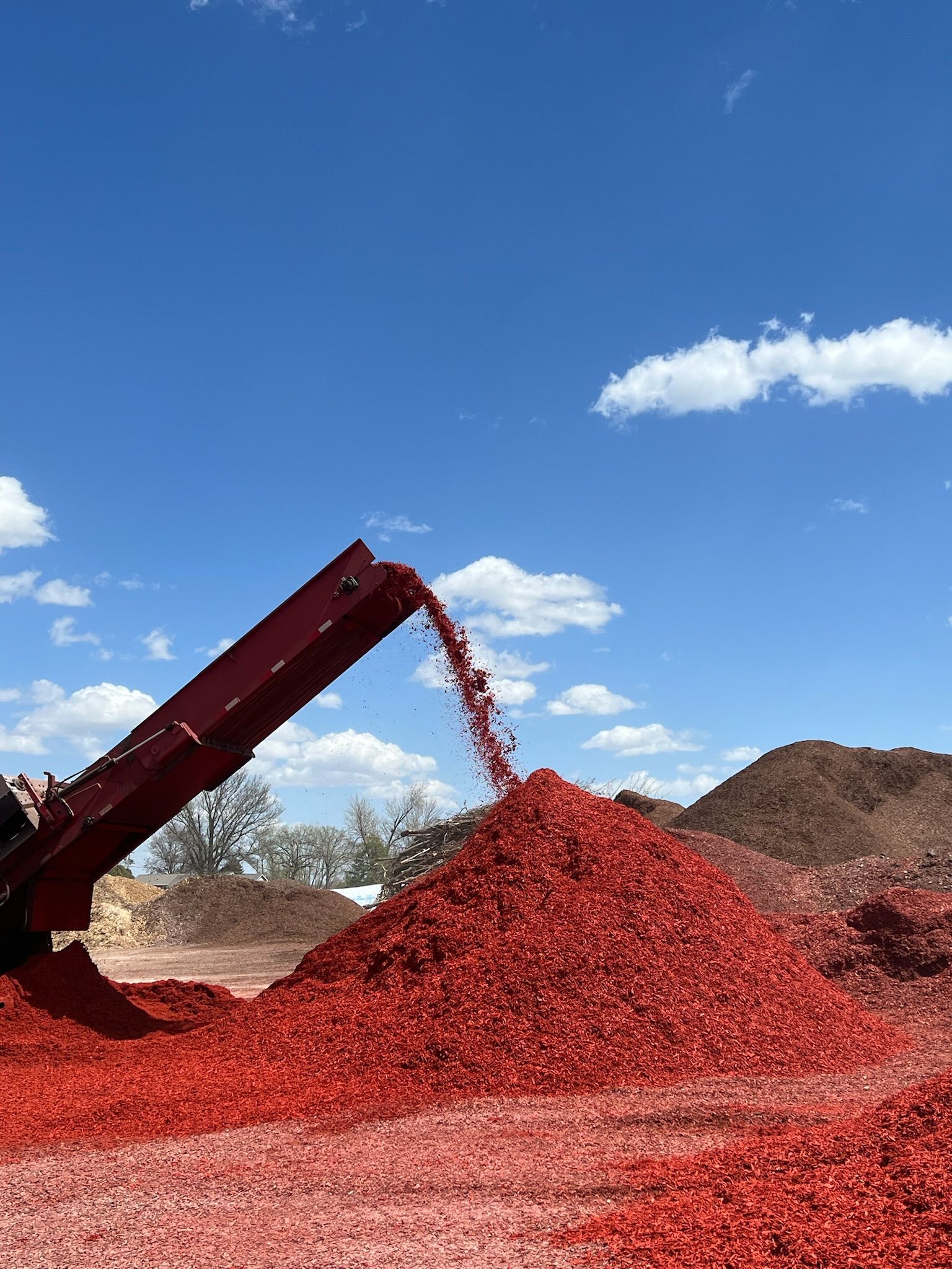Red-colored wood chips pouring from a conveyor belt onto a large pile under a blue sky.