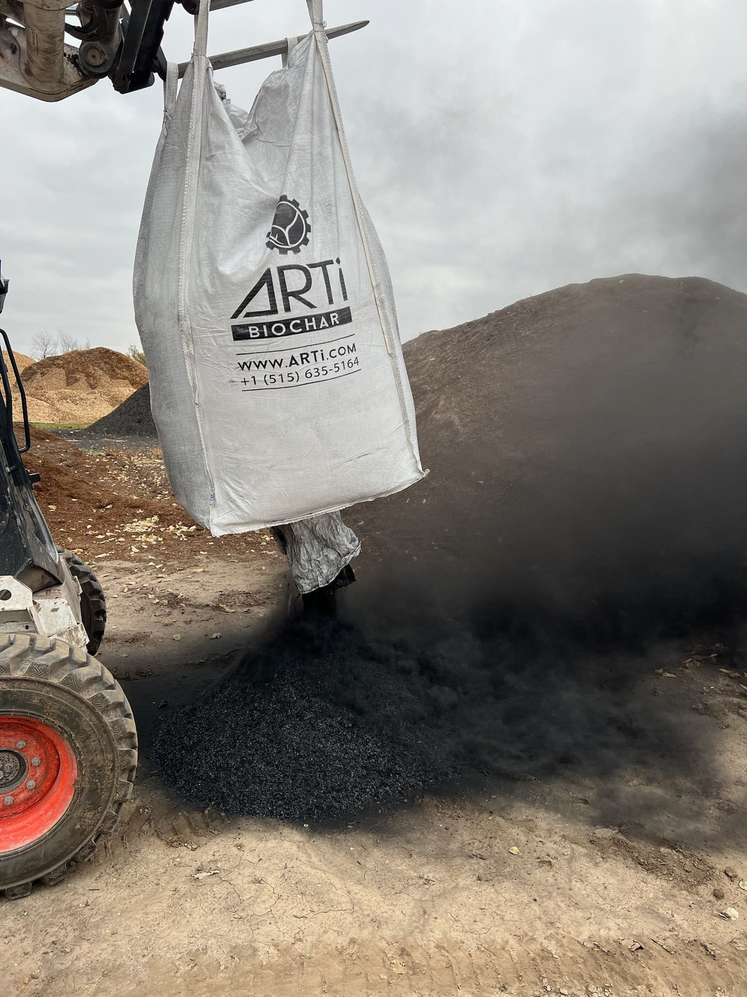 A skid steer dumping a large bag of dark, granular material, likely biochar, onto the ground.