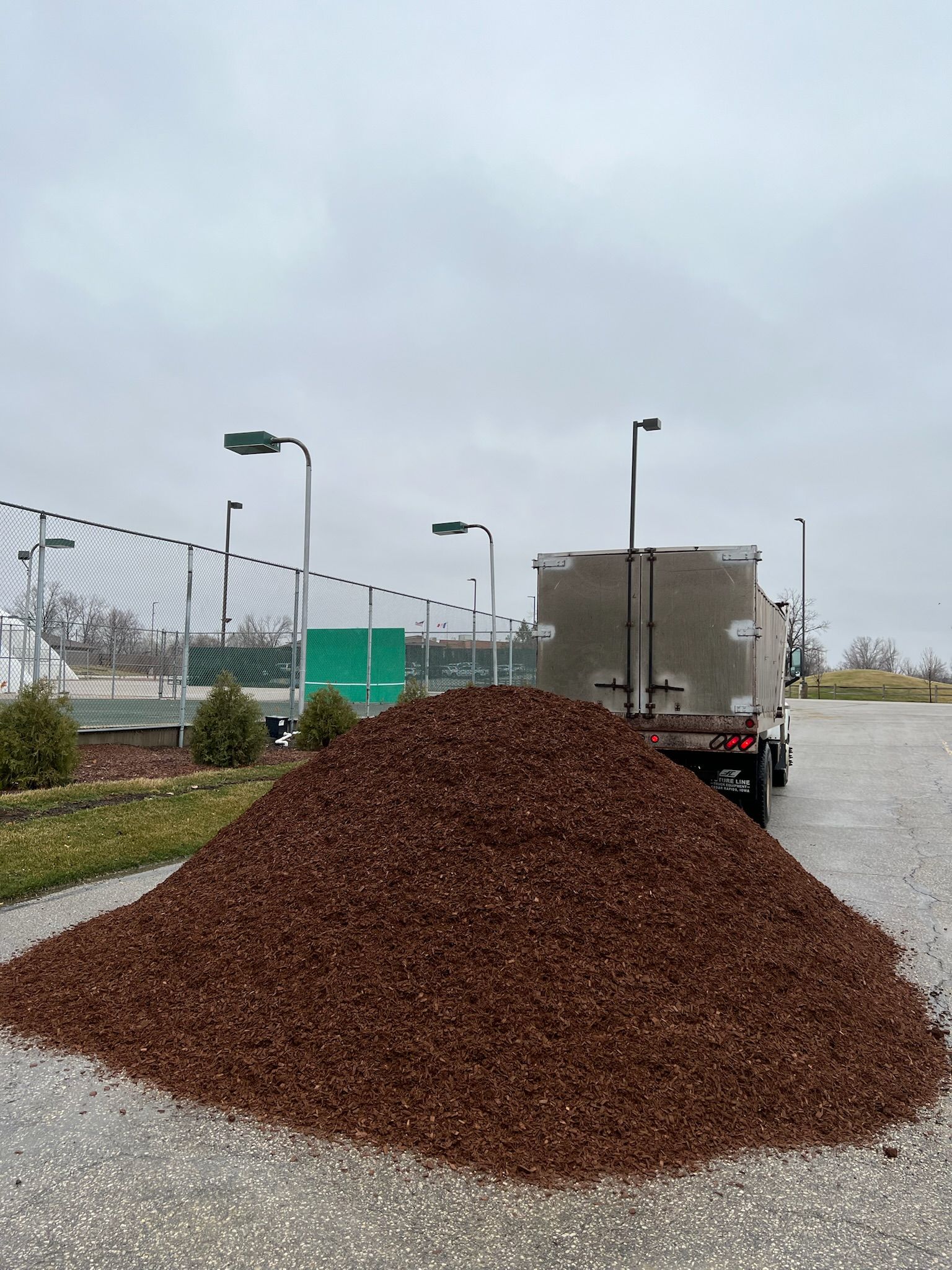 Pile of brown mulch on asphalt in front of a truck, near a fence and under a cloudy sky.