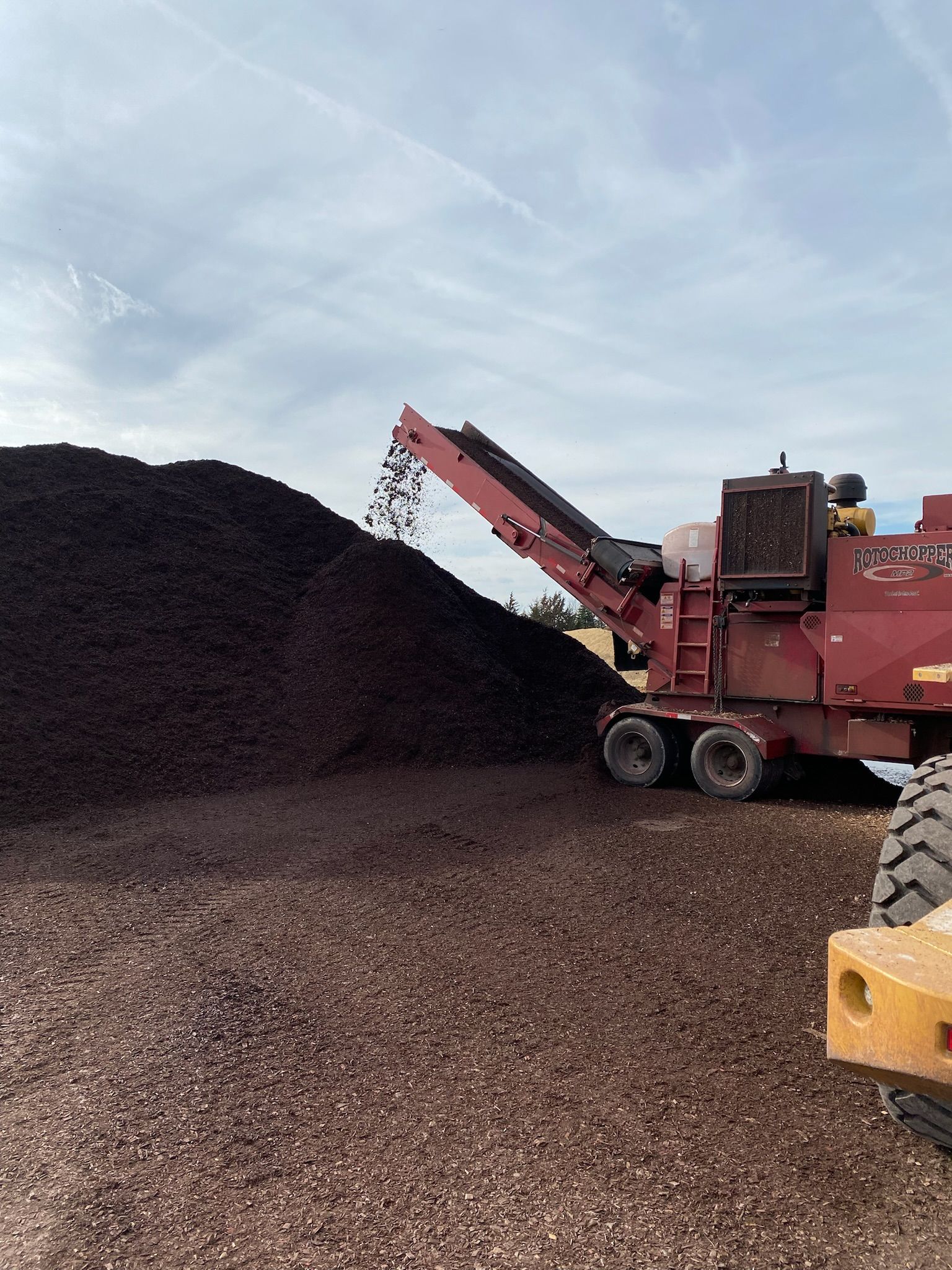Wood chipper depositing wood chips onto a large pile, outdoors, under a cloudy sky.