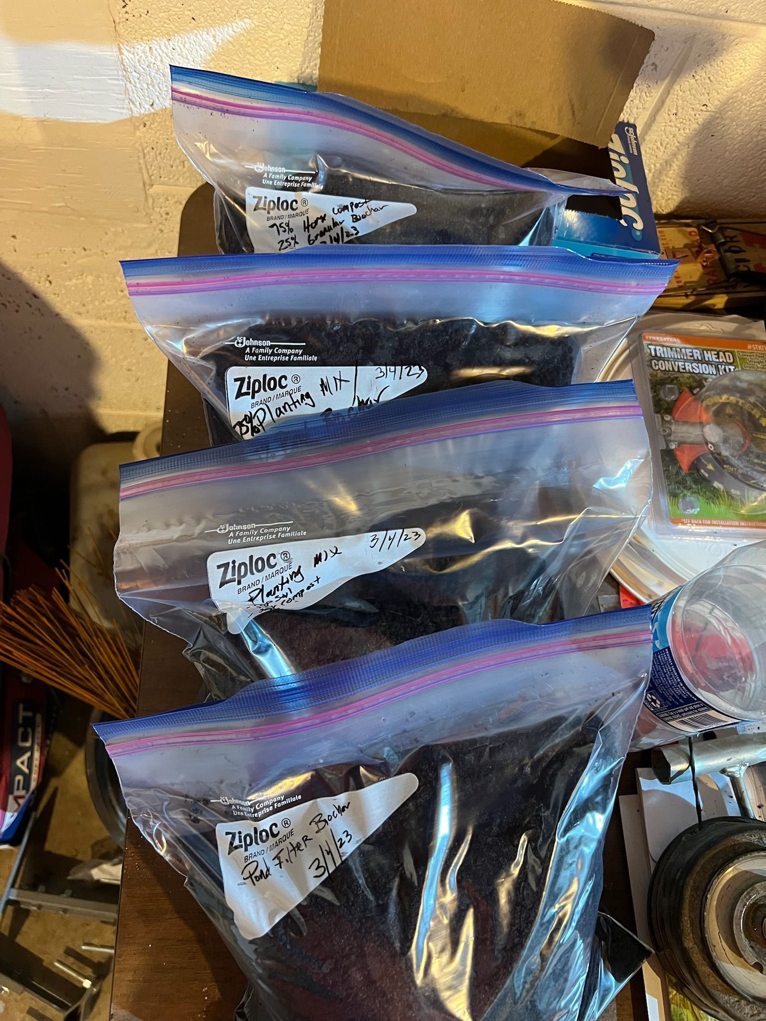 Four clear plastic bags, labeled, filled with dark material, stacked on a wooden surface.
