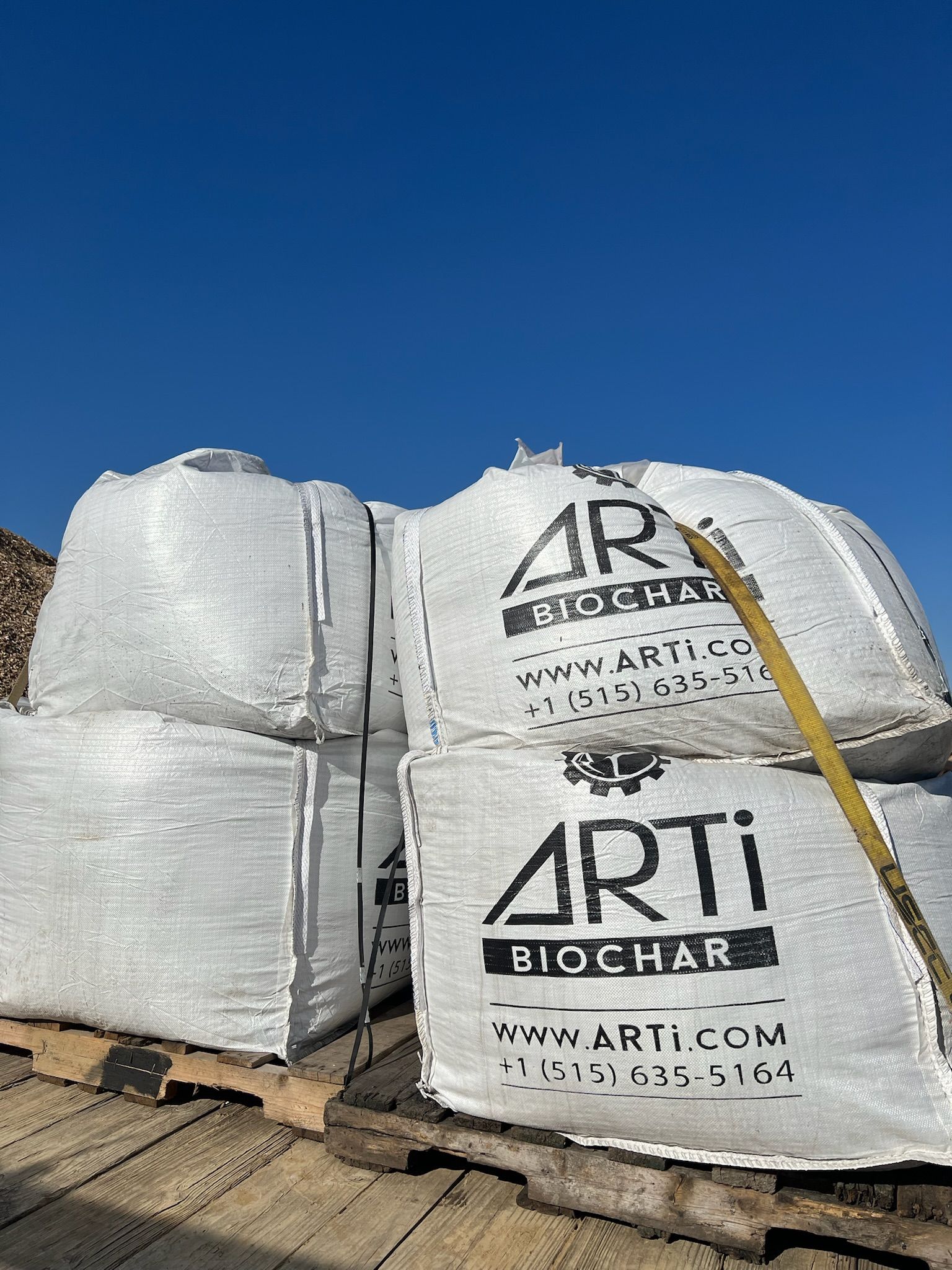 Four white bags of Arti Biochar stacked on a wooden pallet under a blue sky.