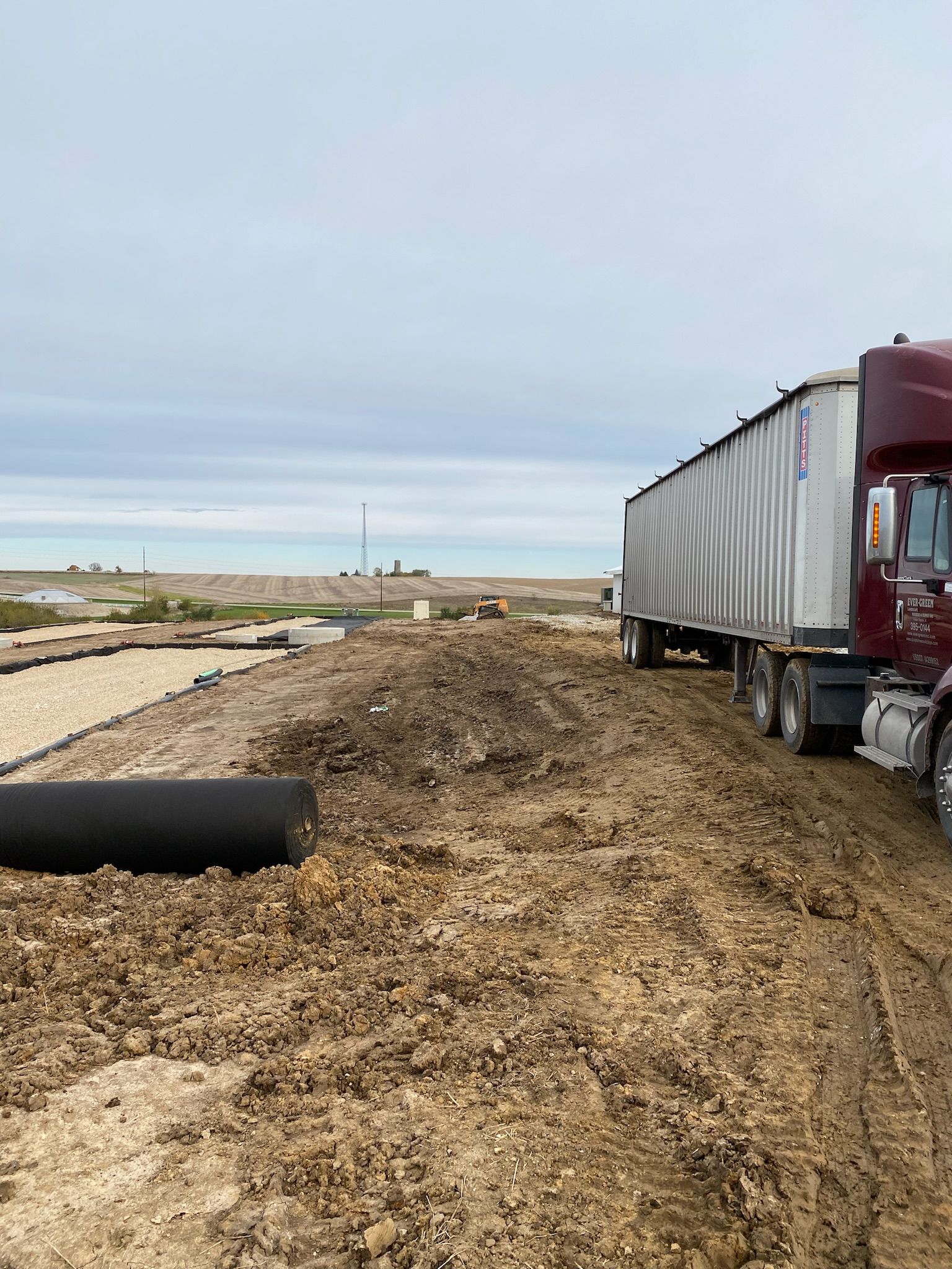 A maroon semi-truck on a dirt road next to a field. Cloudy sky.