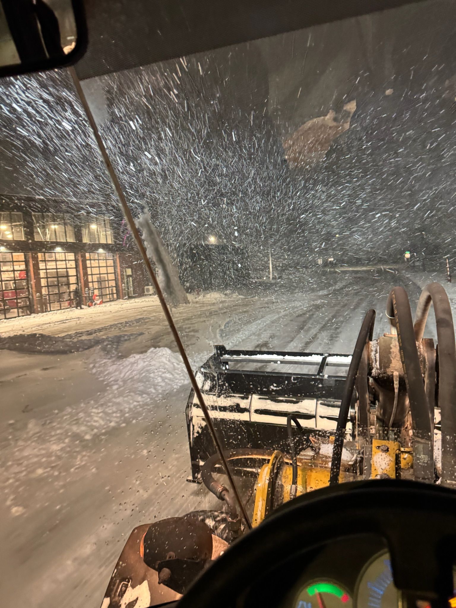 Snowplow clearing a snow-covered road at night, with buildings visible in the background.
