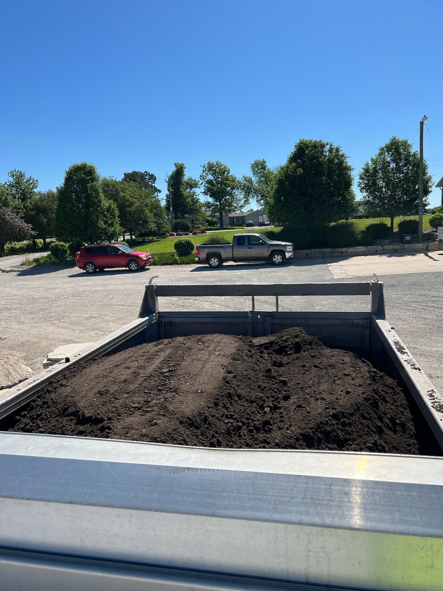 Truck bed filled with dark soil, parked in gravel lot with two vehicles and trees in the background under blue sky.