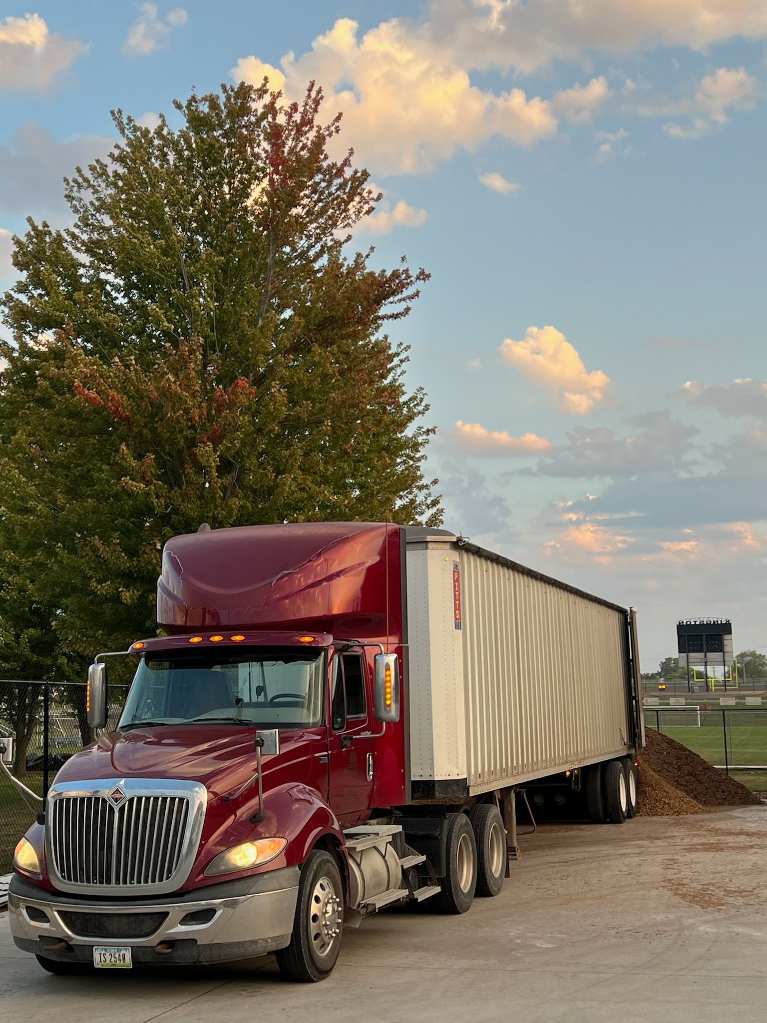 Red semi-truck next to a pile of mulch with a tree and a cloudy sky in the background.