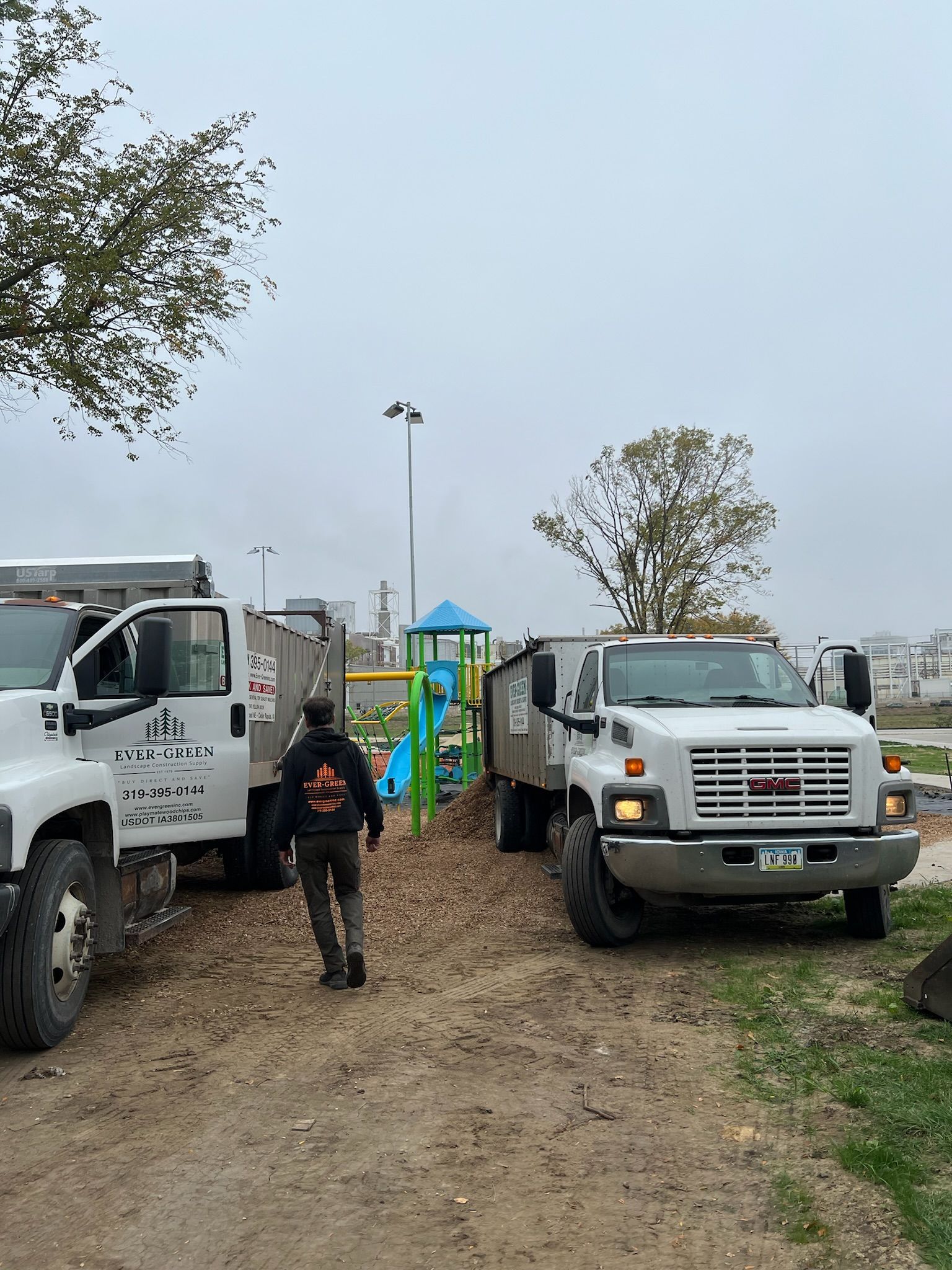 Two dump trucks at a playground; a person walks between them. Grey sky.