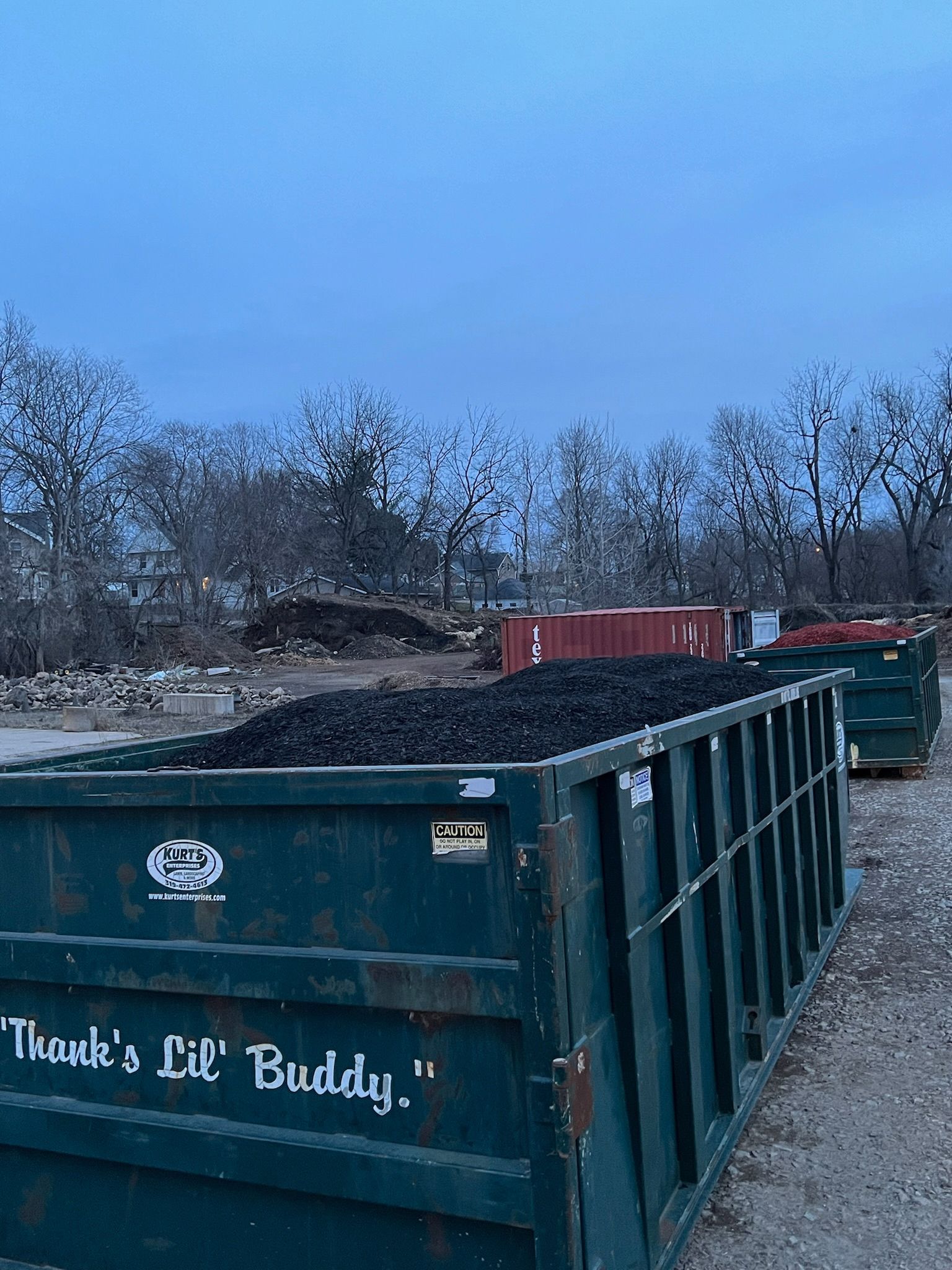 Green dumpsters filled with dark material under a cloudy sky, with bare trees in the background.