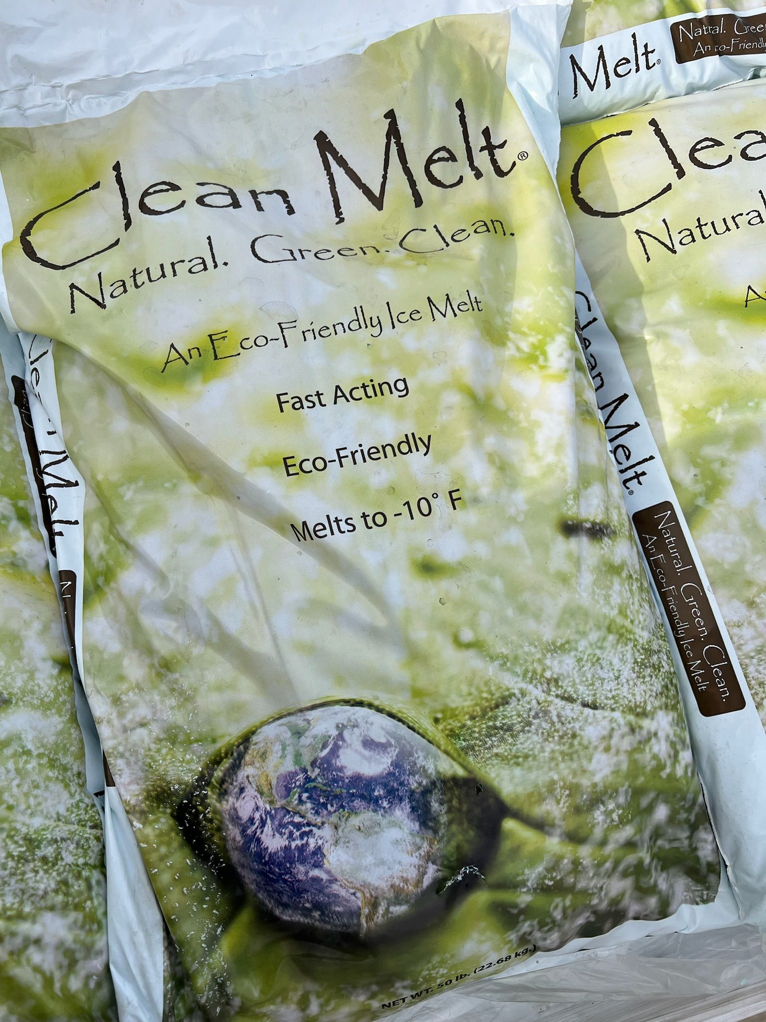 Bags of Clean Melt natural de-icer. White bags with green and brown text, earth graphic, 
