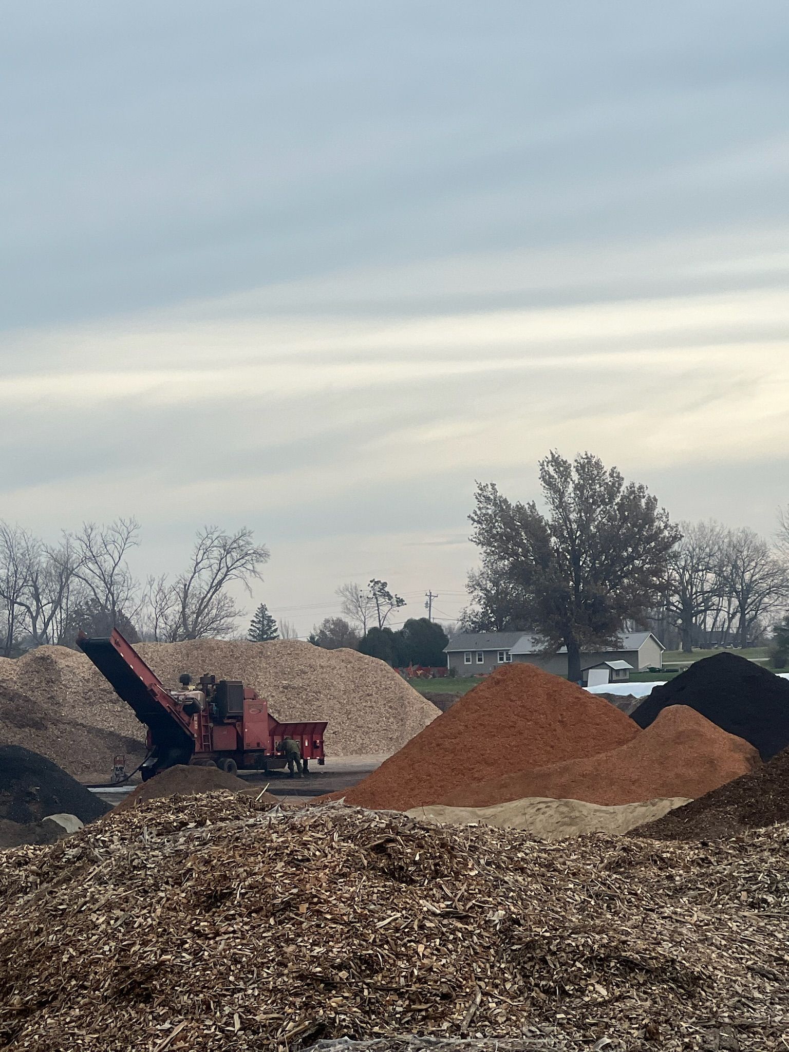 Mulch piles of various colors with a wood chipper in an outdoor setting under a cloudy sky.
