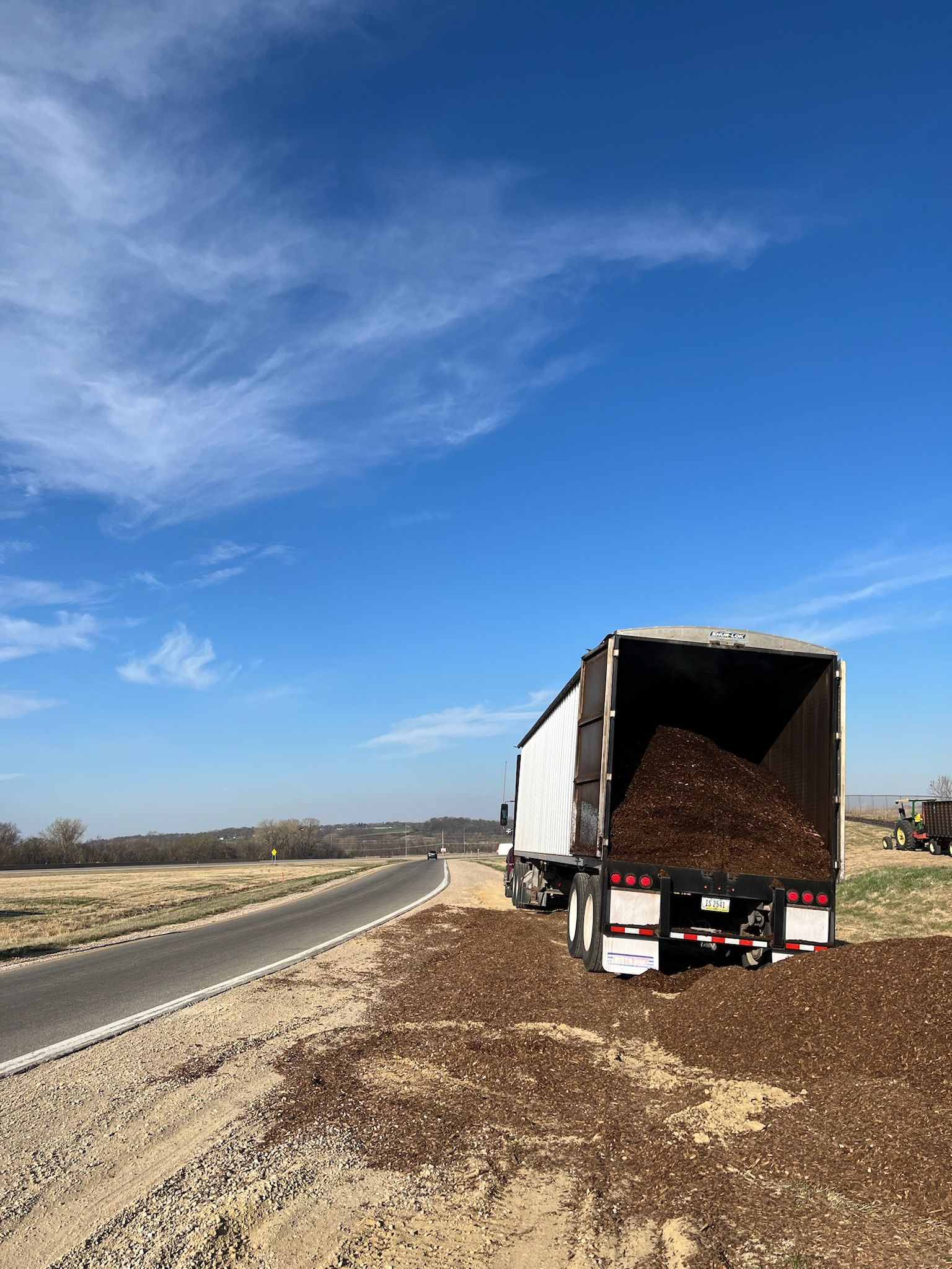 Semi-truck unloading mulch onto a dirt shoulder next to a paved road under a blue sky.