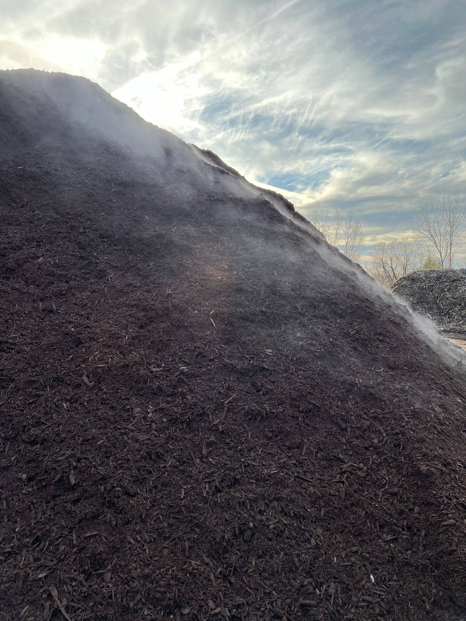 Large pile of dark mulch steaming under a partly cloudy sky.