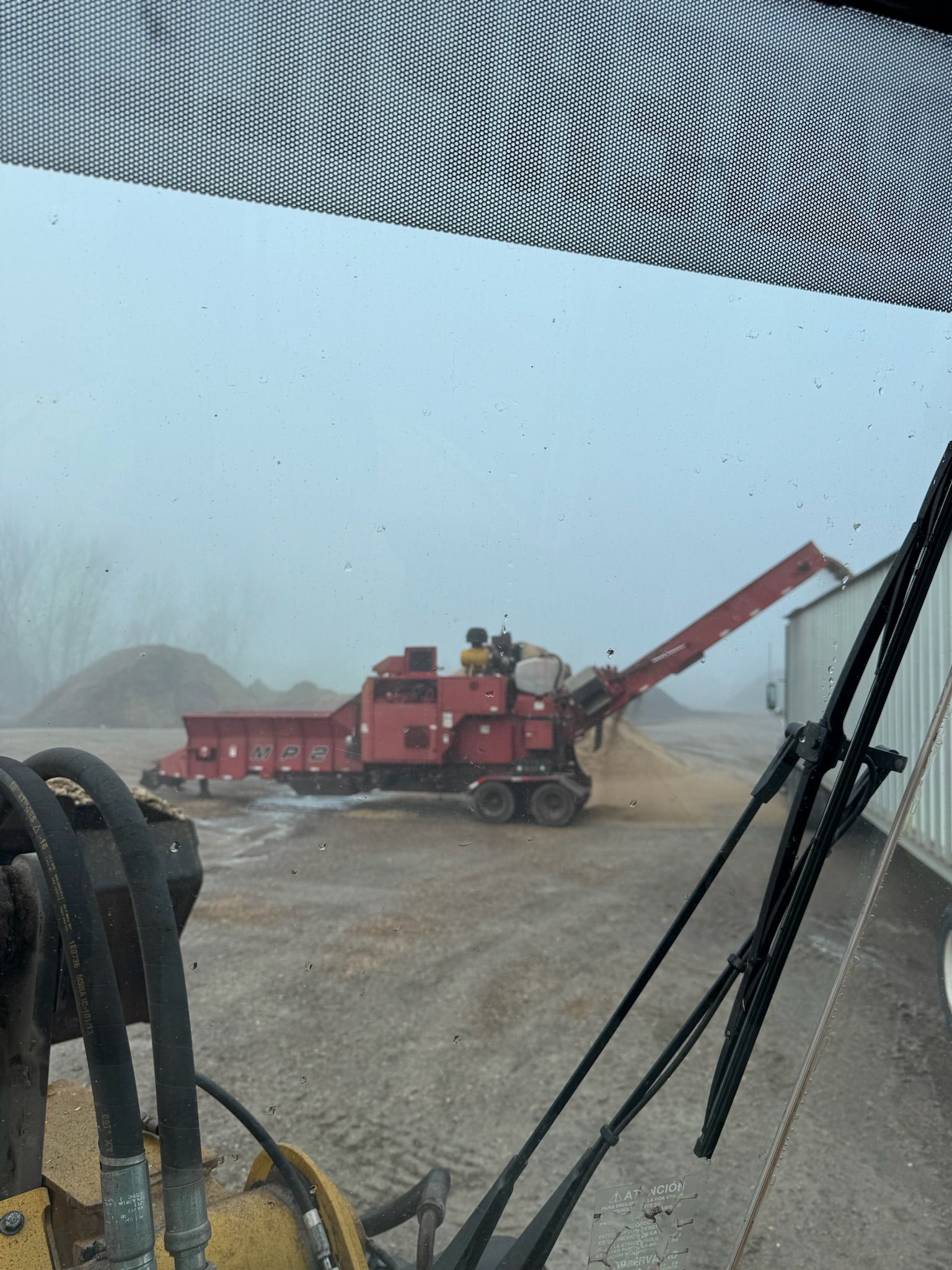 A red industrial machine with a conveyor belt is unloading material near a building on a foggy day.