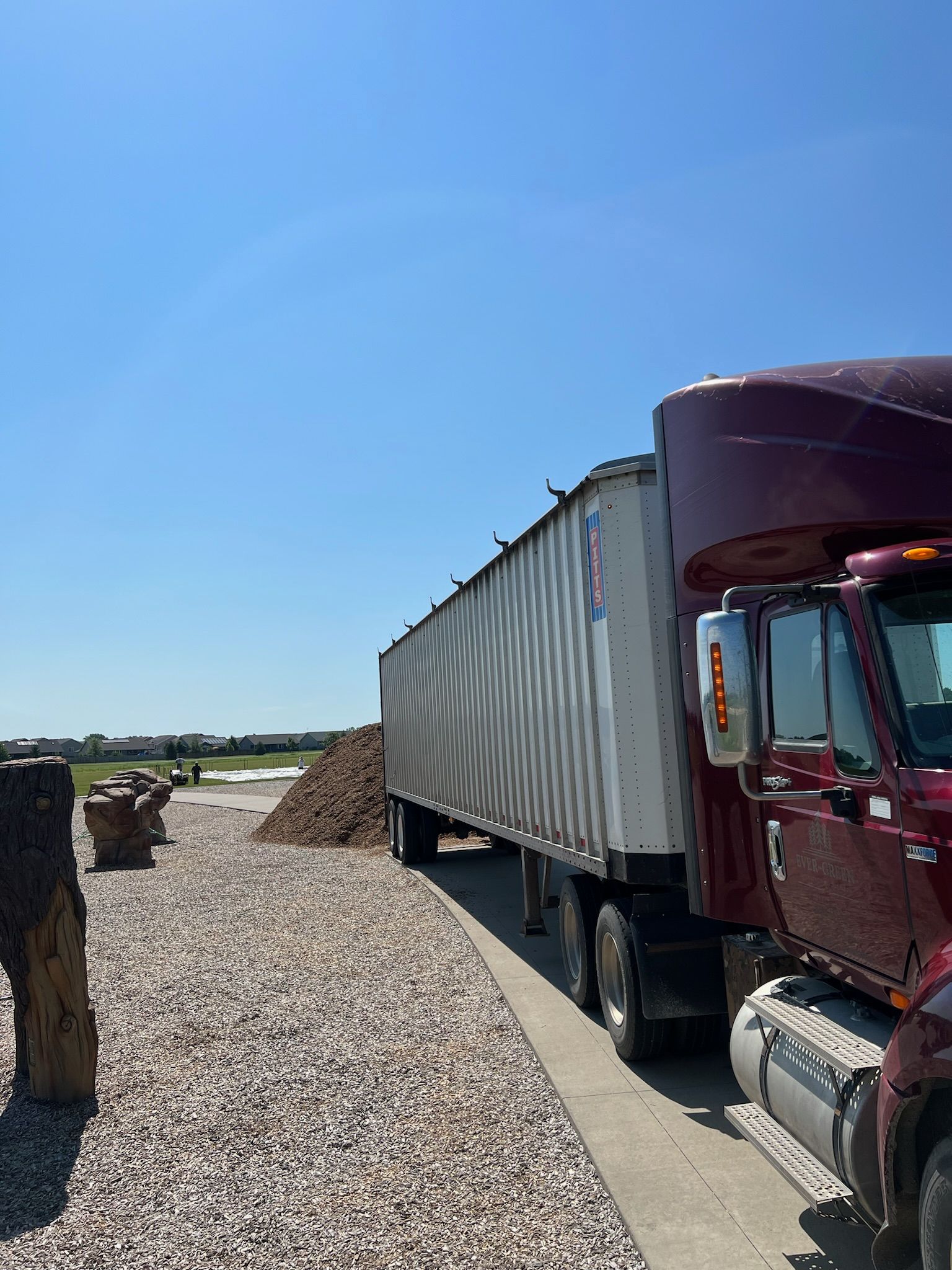 Red semi-truck next to a gravel-covered area. Its trailer is filled with mulch. Blue sky overhead.