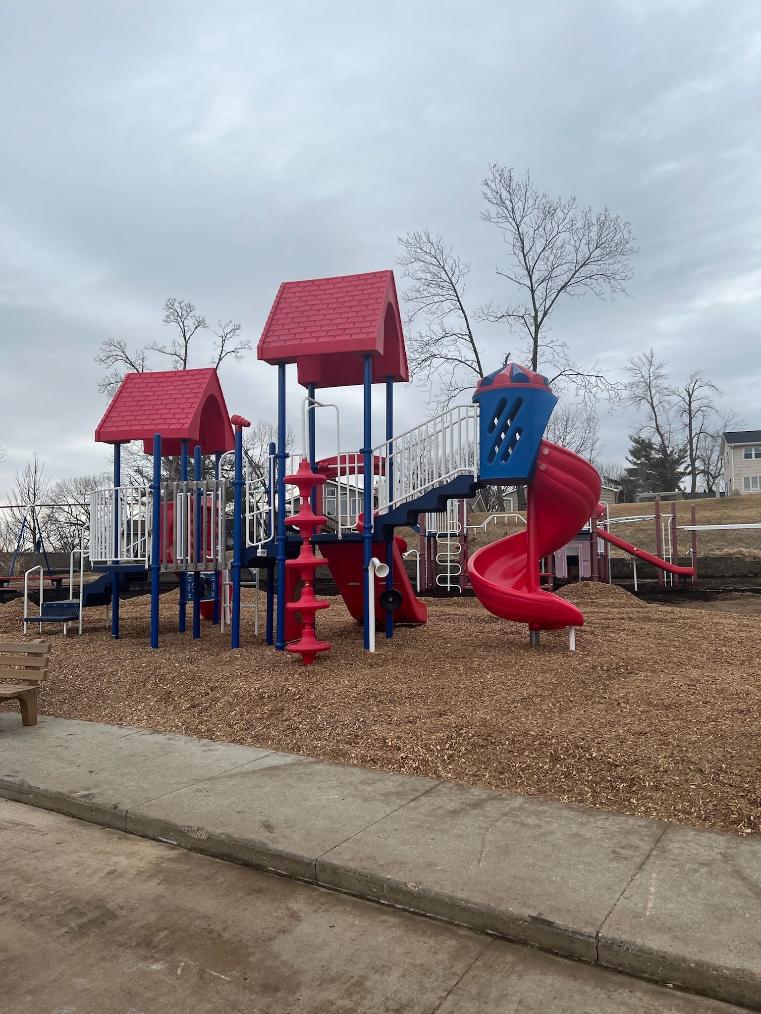 Playground with red and blue play structures, wood chip surface, and cloudy sky.