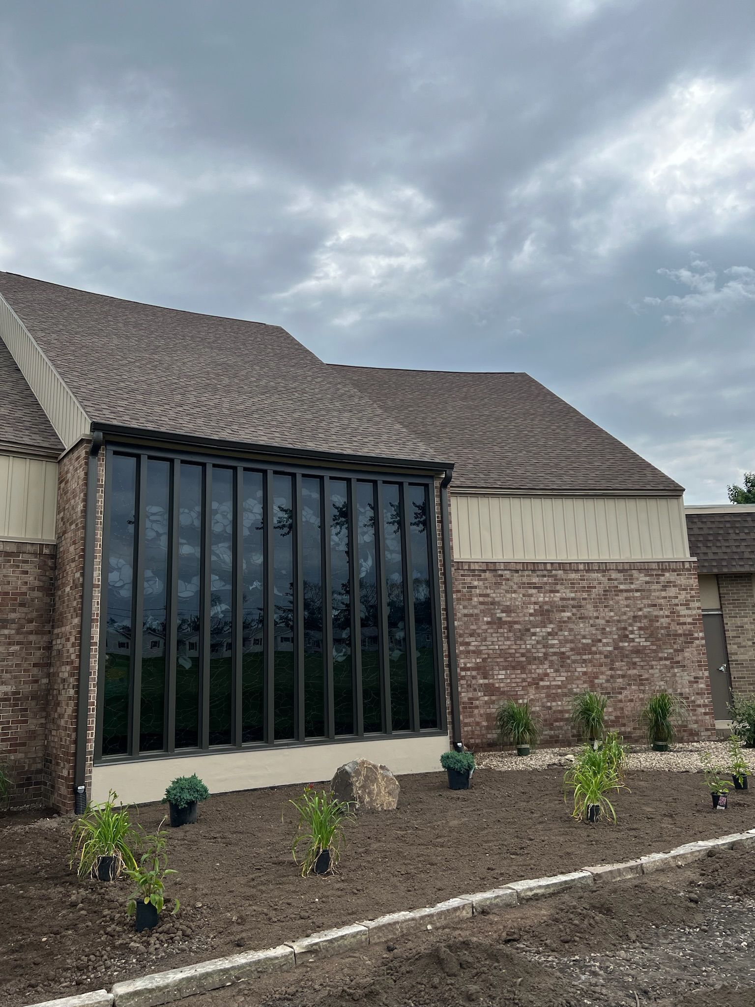 Brick building with dark stained windows, brown roof, and freshly planted garden. Cloudy sky.