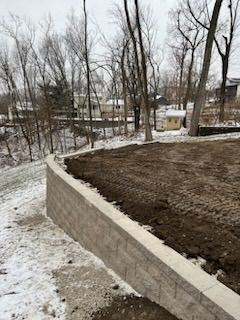 A retaining wall on a hillside with freshly turned soil, trees in the background, and snow on the ground.