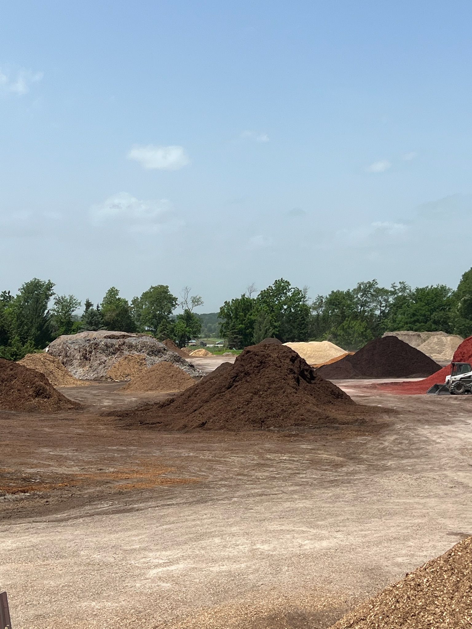 Piles of mulch in different colors, set on a gravel lot, with trees and sky in the background.