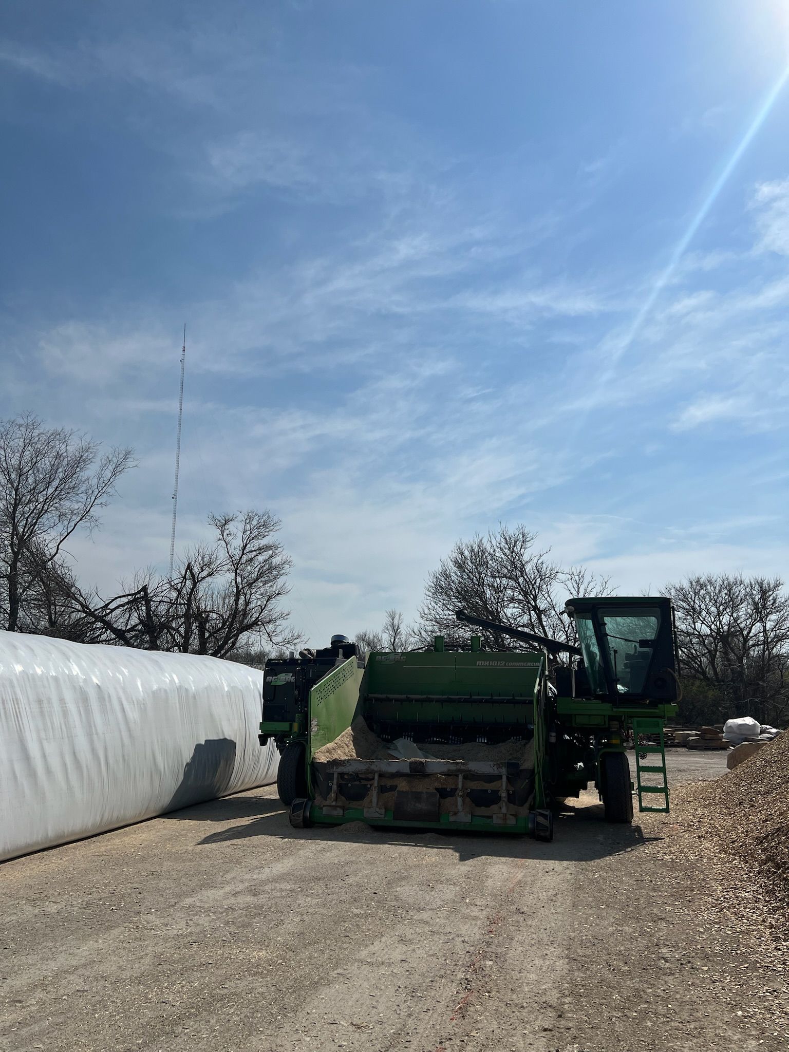 Green forage harvester next to a large white silage storage bag on a gravel lot under a blue sky.