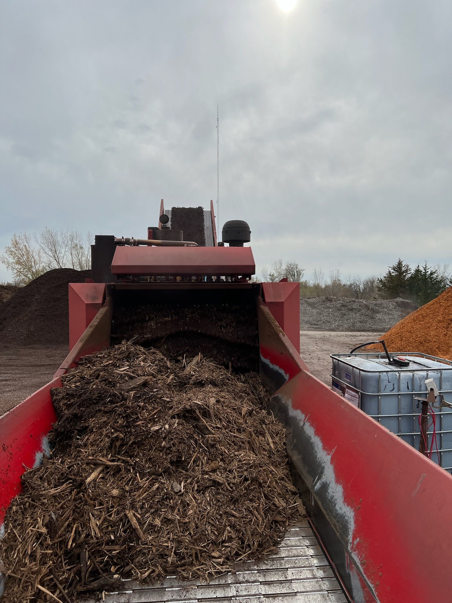 A red industrial machine overflowing with wood chips, outdoors on a cloudy day.