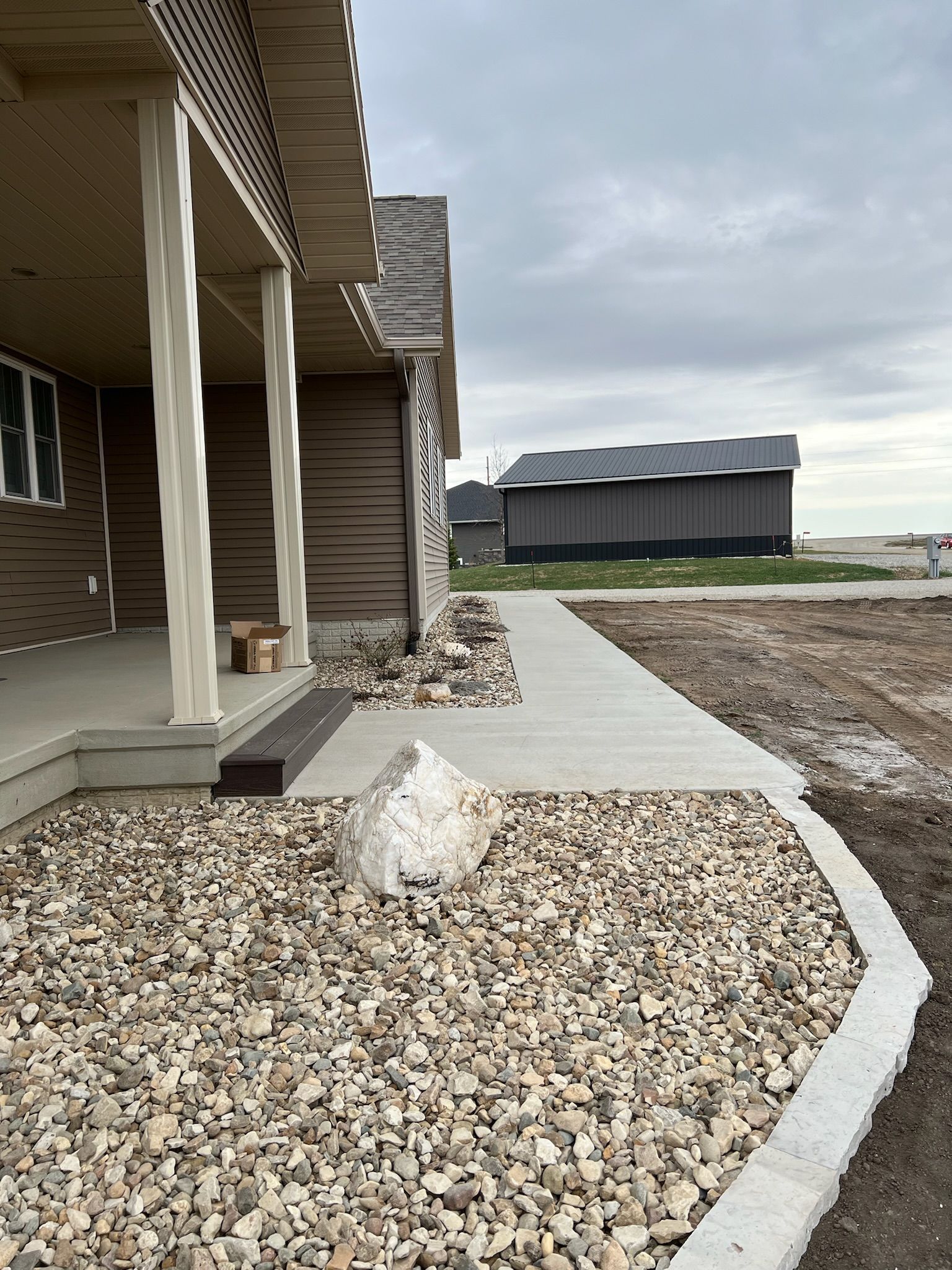 House with a covered porch and gravel landscaping; a concrete walkway extends toward a distant building on a cloudy day.