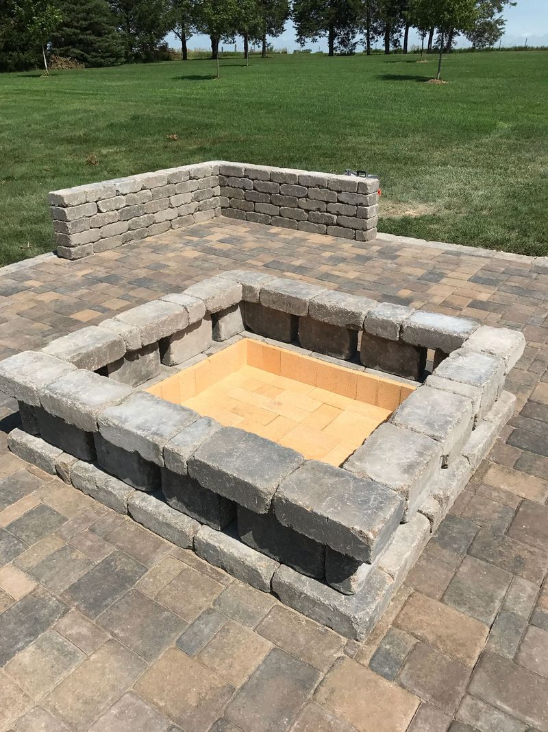 Square fire pit built with gray stone blocks, on a brick patio. Back wall in background.