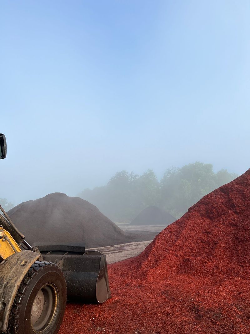 A yellow front-end loader next to mounds of red mulch, with a foggy background.