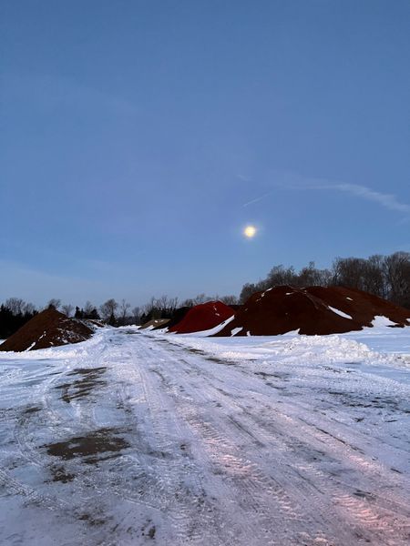 Snow-covered dirt road with red soil piles and trees under a light blue sky with a bright sun.