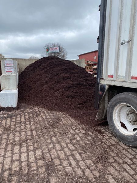 Large pile of dark mulch beside a truck on a brick paved area, under a cloudy sky.