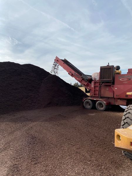 A red wood chipper deposits wood chips onto a large pile on a brown gravel surface under a cloudy sky.
